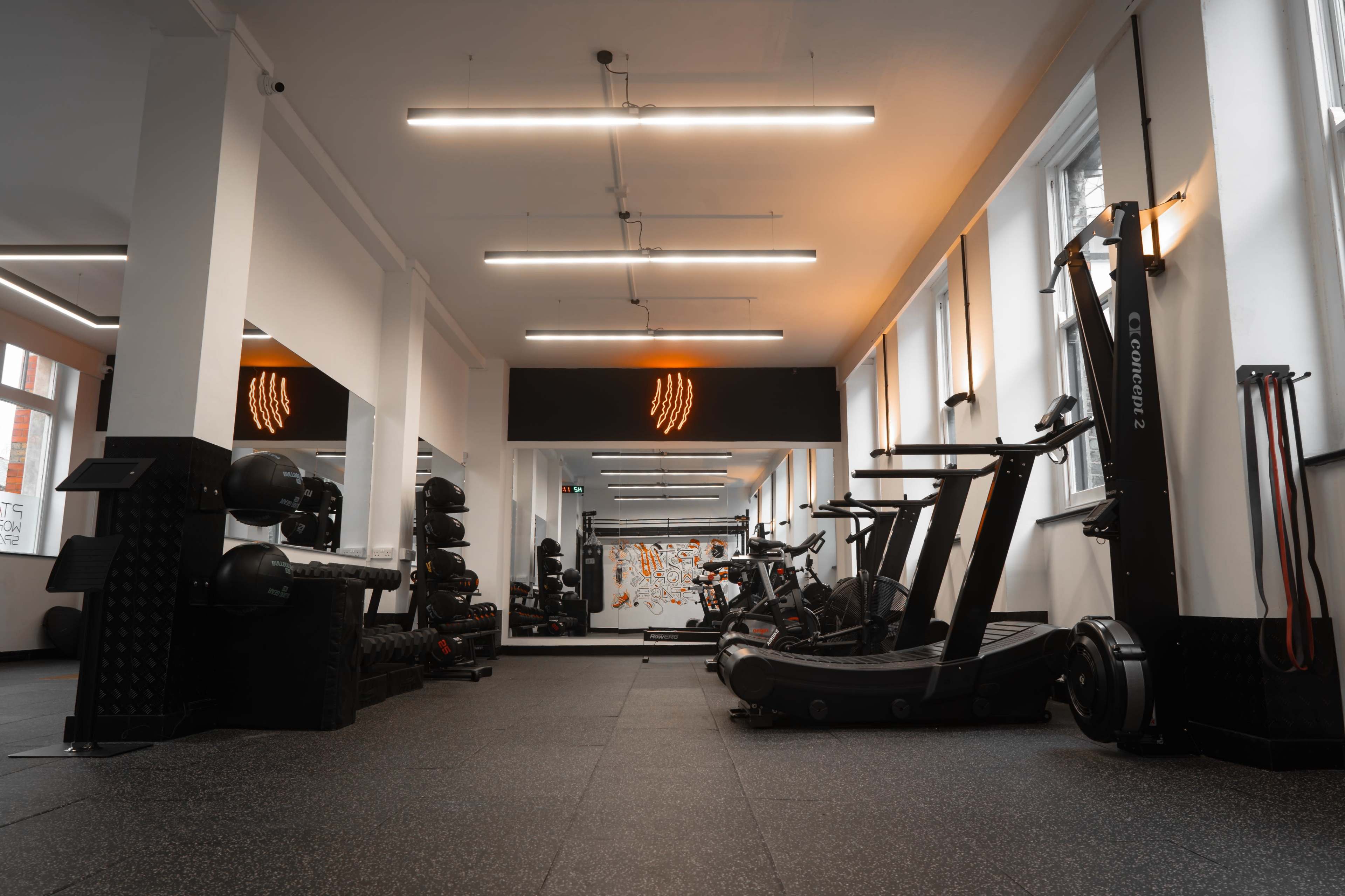 The image shows a modern gym with various fitness equipment against a backdrop of bright lighting and large mirrors.