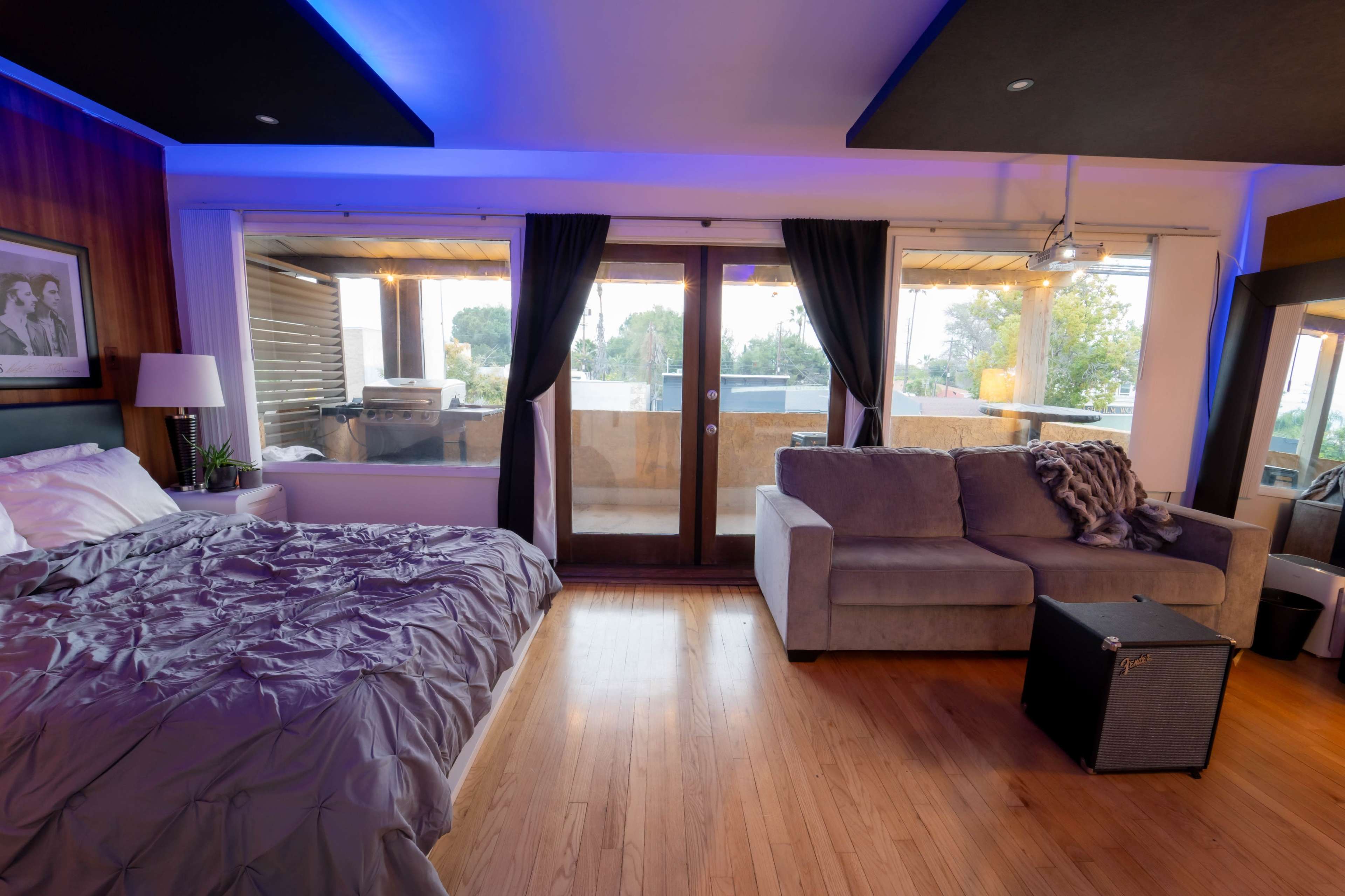 A modern bedroom features a bed with gray linens, a couch, and large windows that open to a balcony.