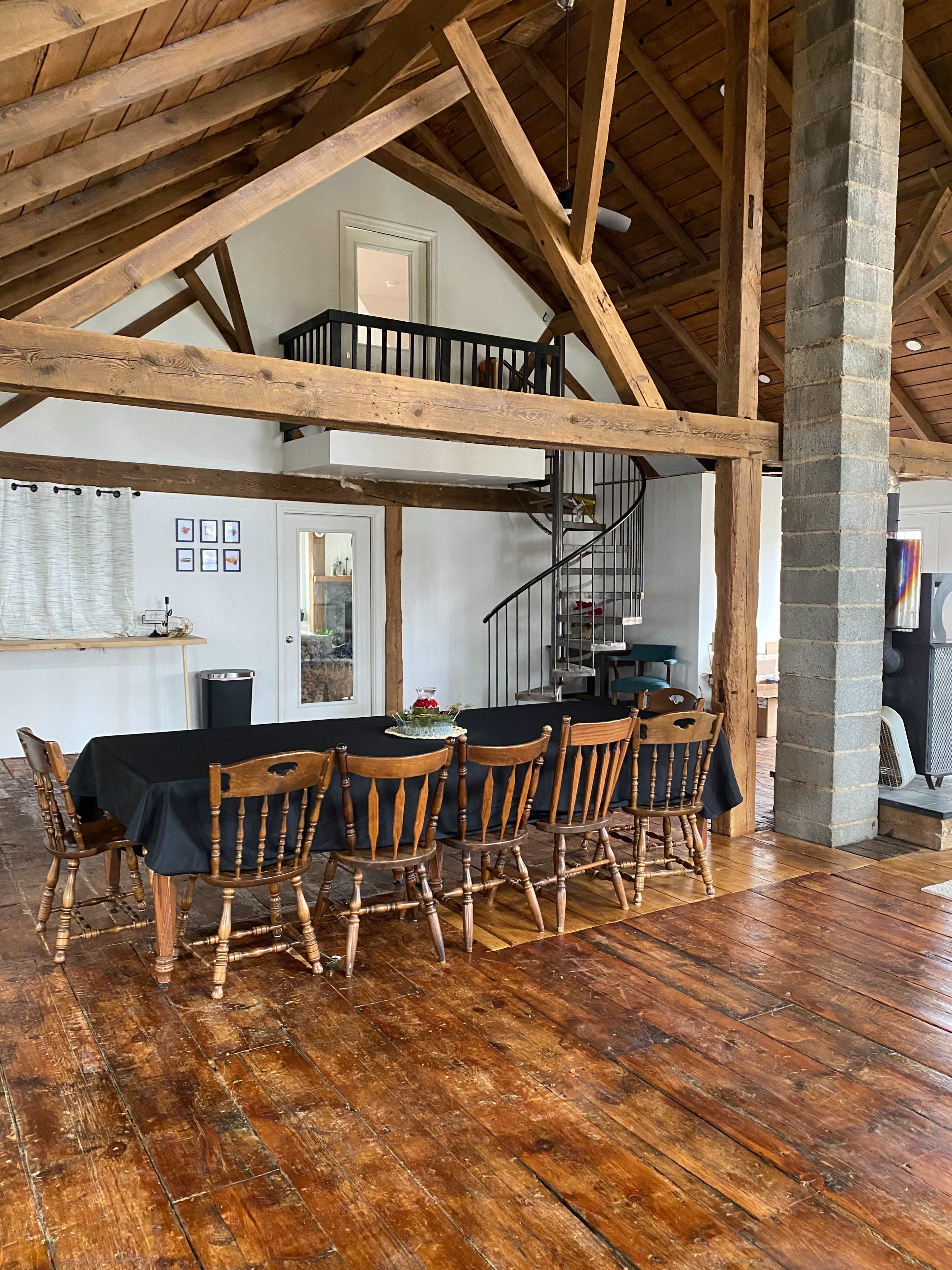 A large dining table with wooden chairs is set in a spacious room featuring exposed beams, a spiral staircase, and a mix of modern and rustic decor.
