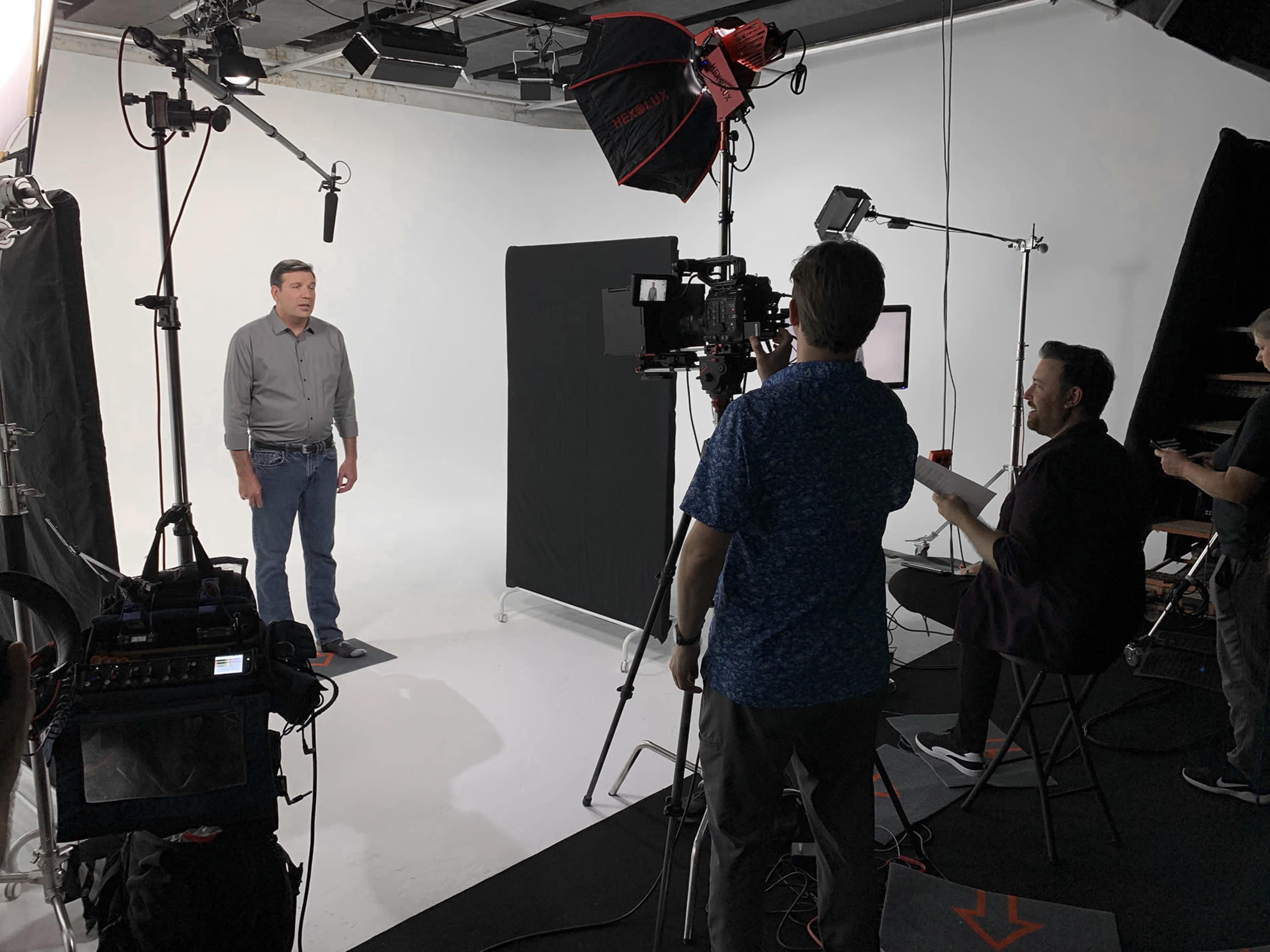 A man stands in front of a camera setup on a simple studio set, while a crew operates equipment in the background.