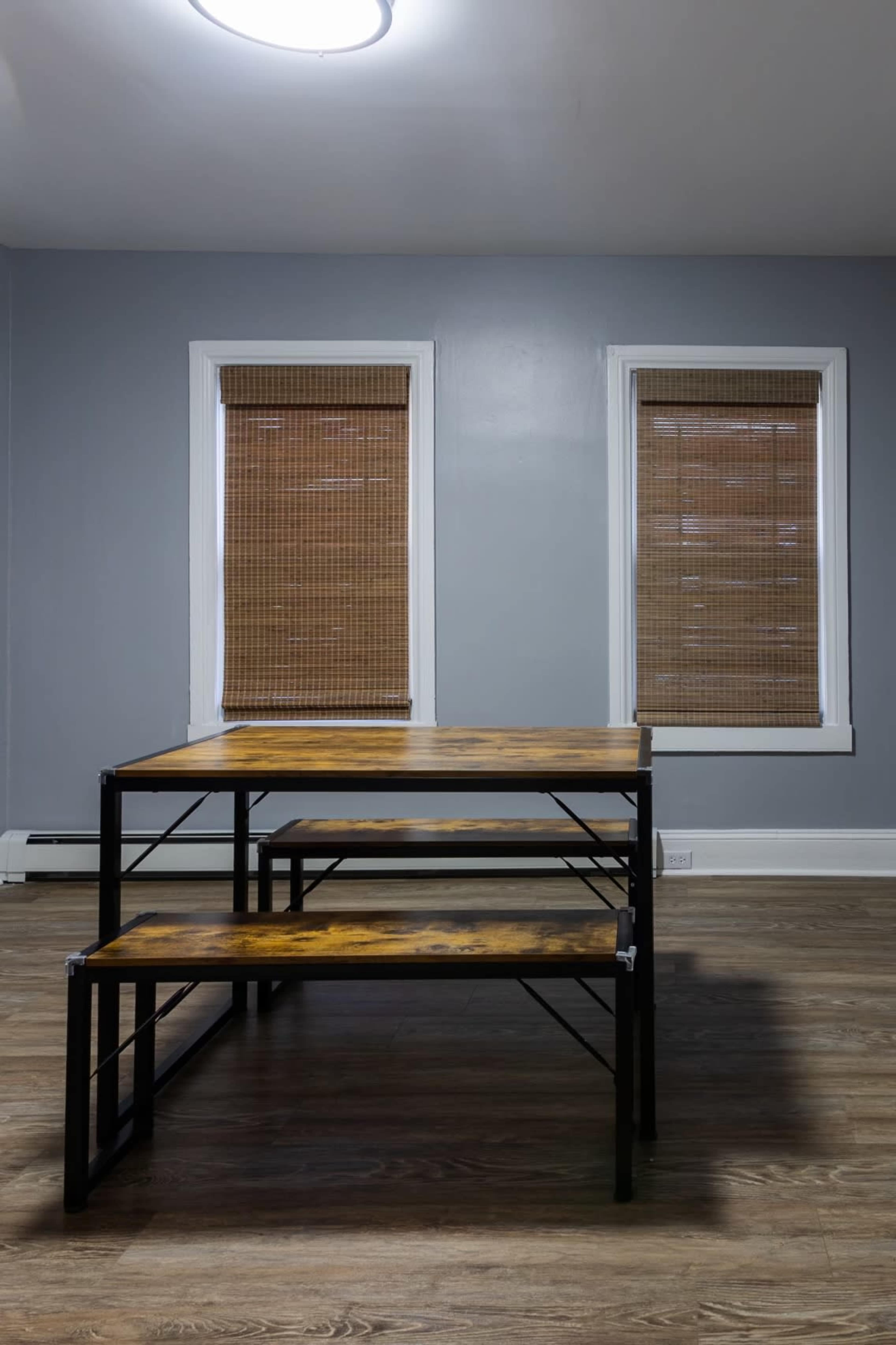 A wooden table with metal legs is positioned in the center of a room with gray walls and two windows covered by bamboo blinds.