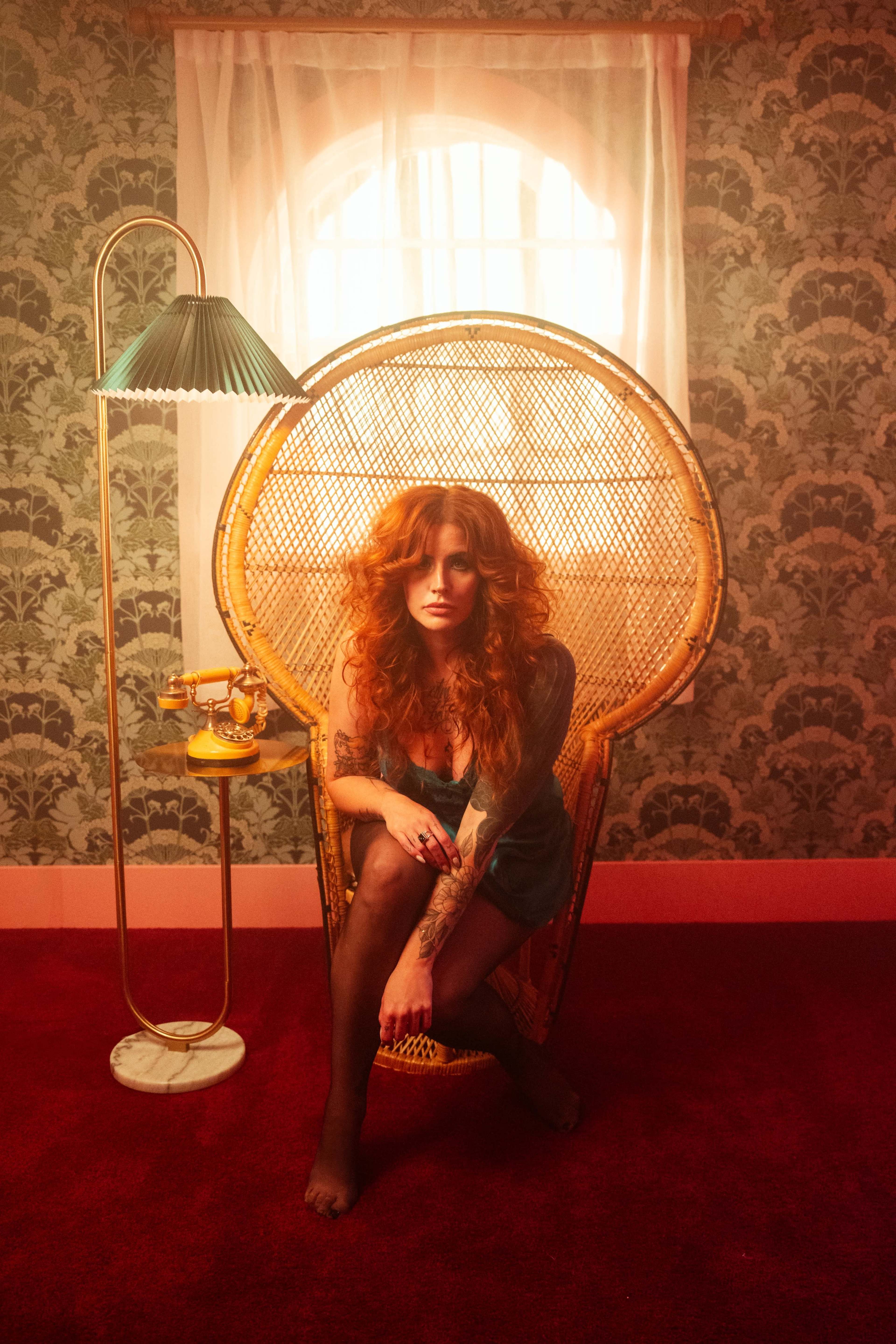 A woman with long, curly red hair sits in a large rattan chair, framed by a softly lit room with patterned wallpaper and a floor lamp beside her.