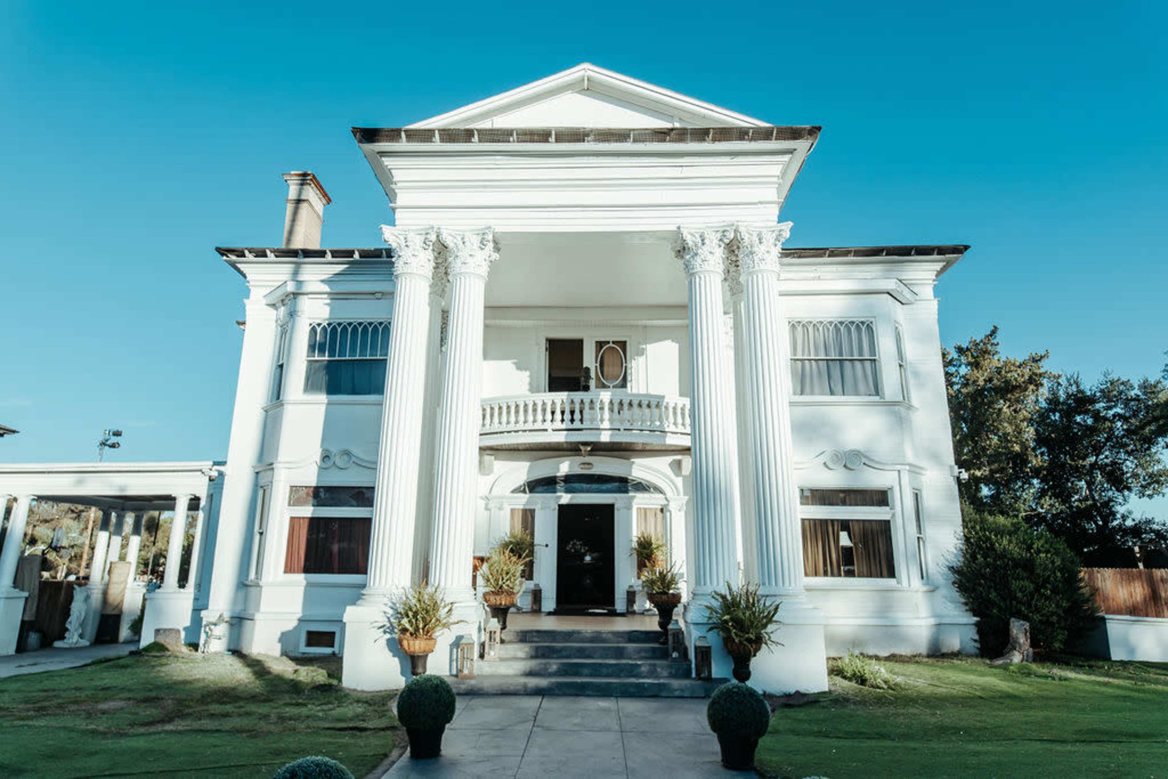 The image shows a large, white, colonial-style mansion featuring four tall columns and a wide staircase leading to the entrance.