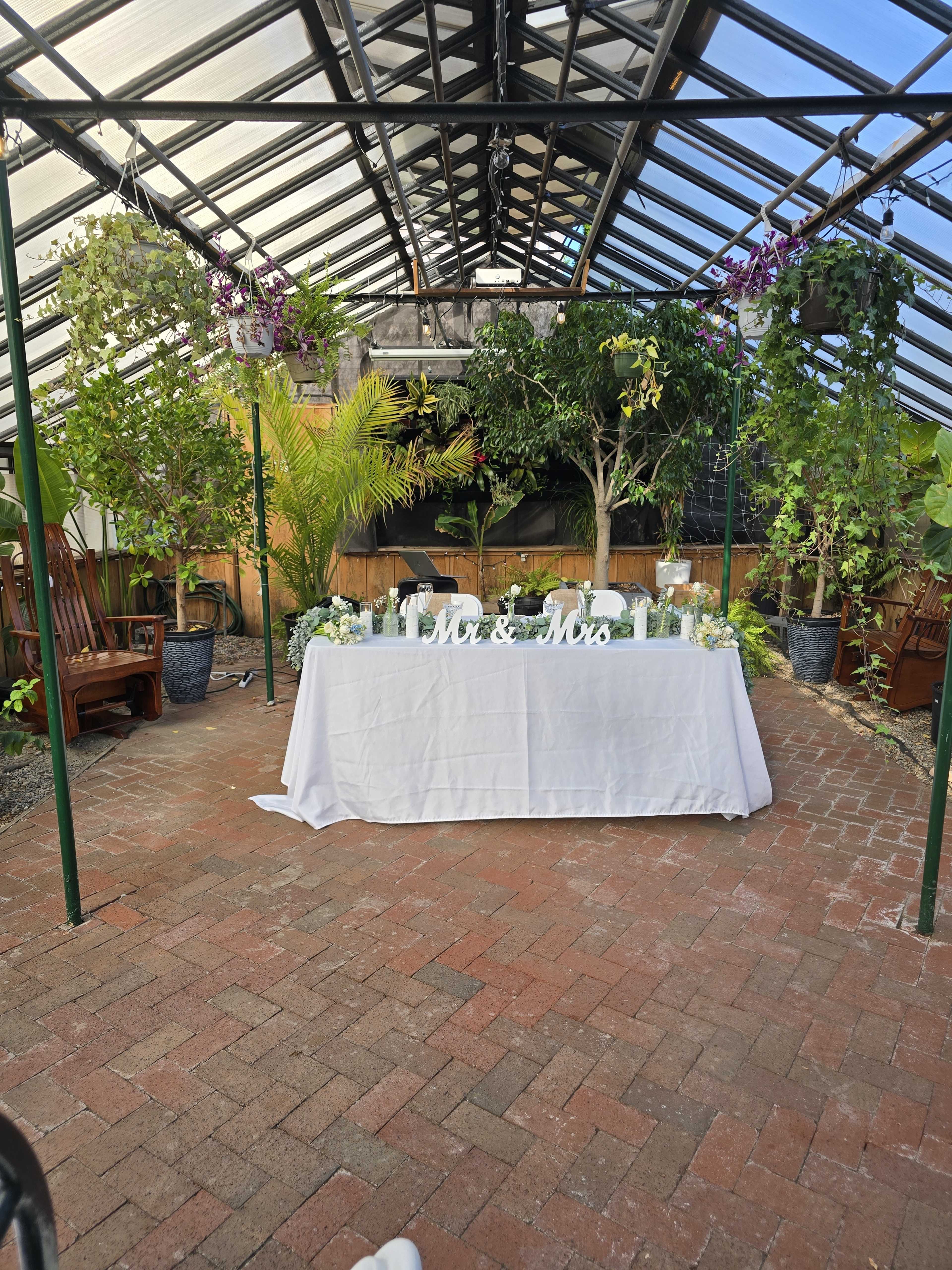 A decorated table with the words "Mr. & Mrs." is set up in a greenhouse surrounded by plants and flowers.
