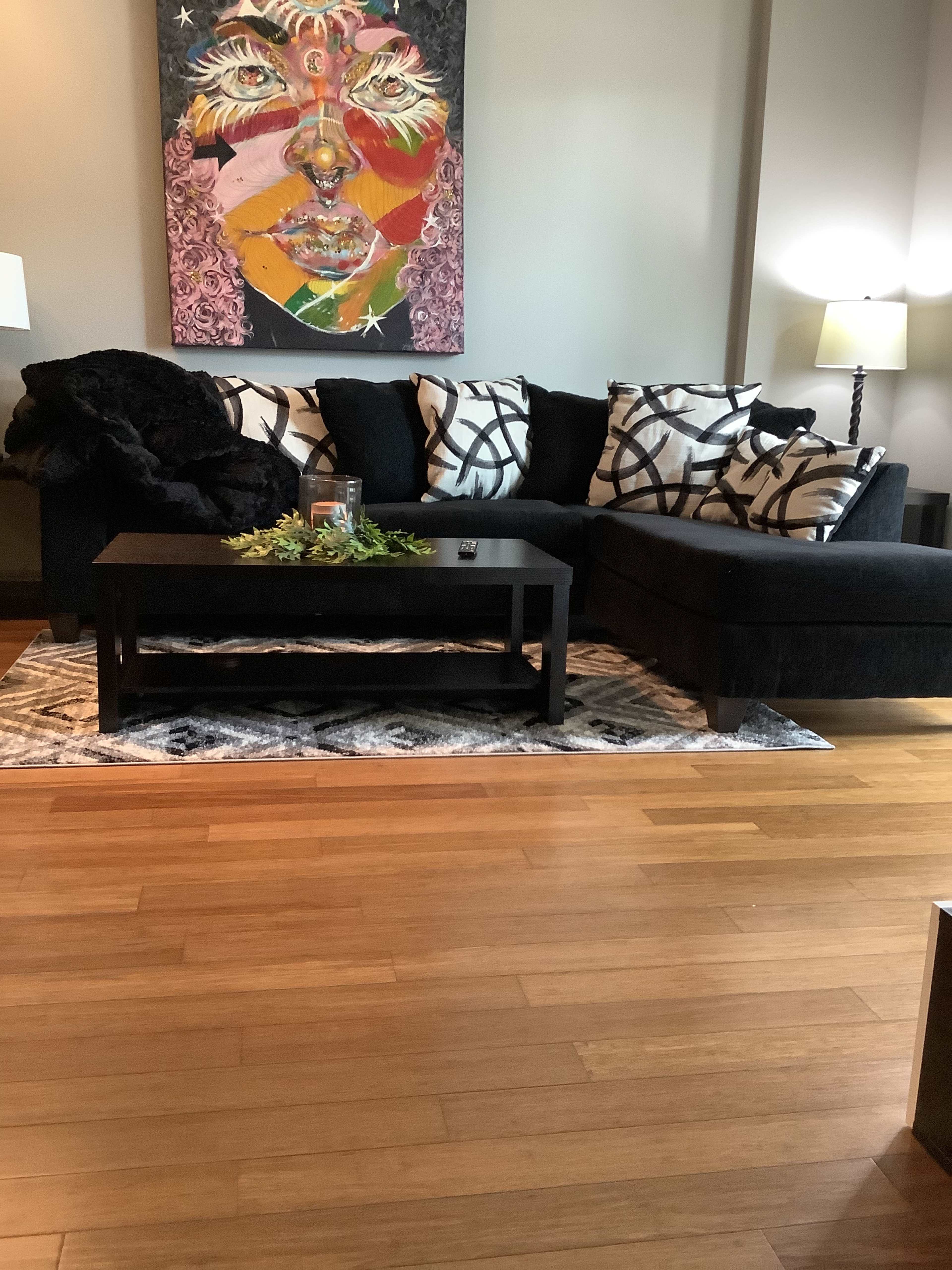 A sectional black sofa adorned with patterned cushions, a coffee table on a decorative rug, and a wall with an abstract portrait painting.