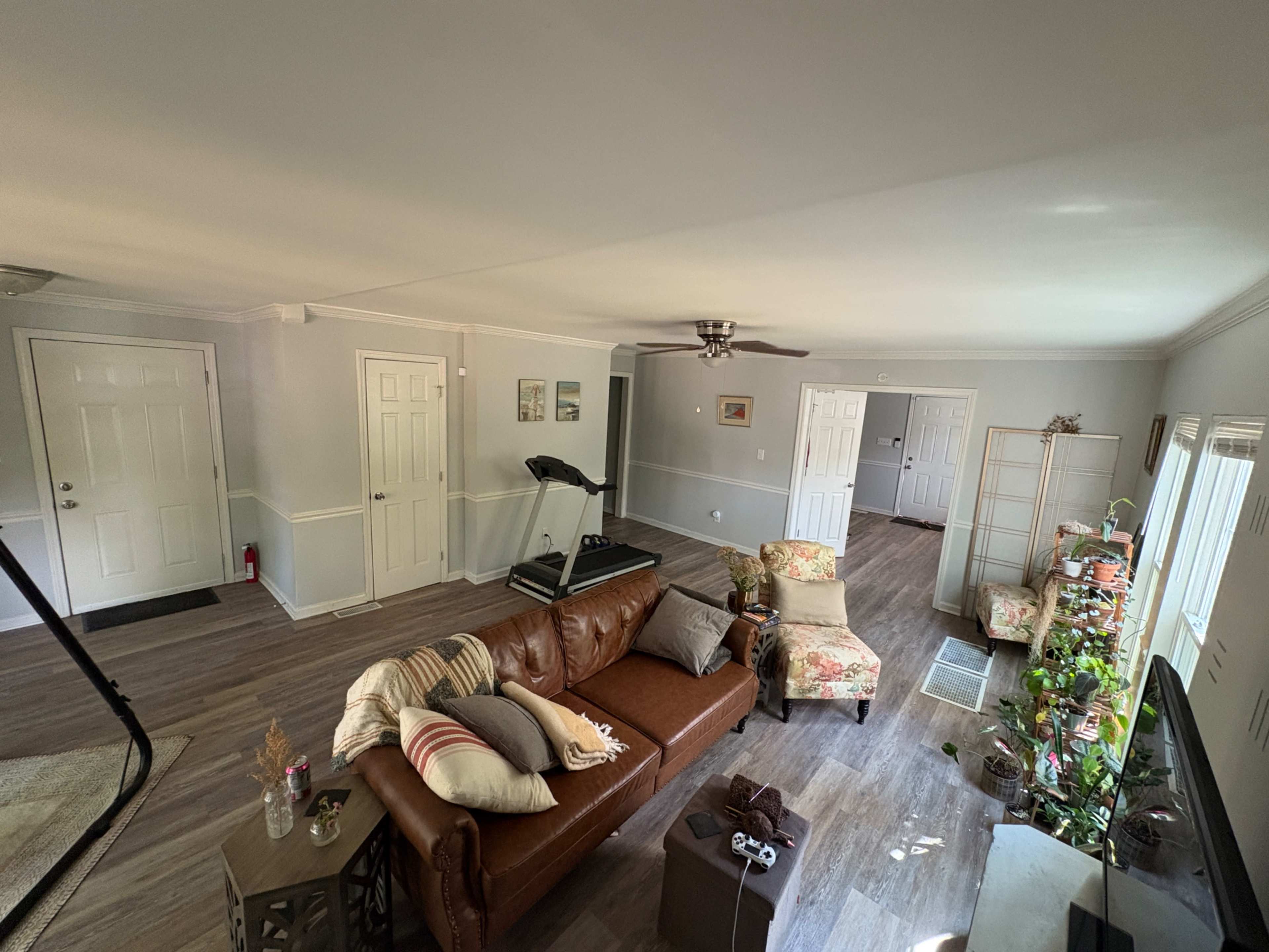 The living area features a brown leather sofa, a treadmill, and various plants, with light-colored walls and wooden flooring.