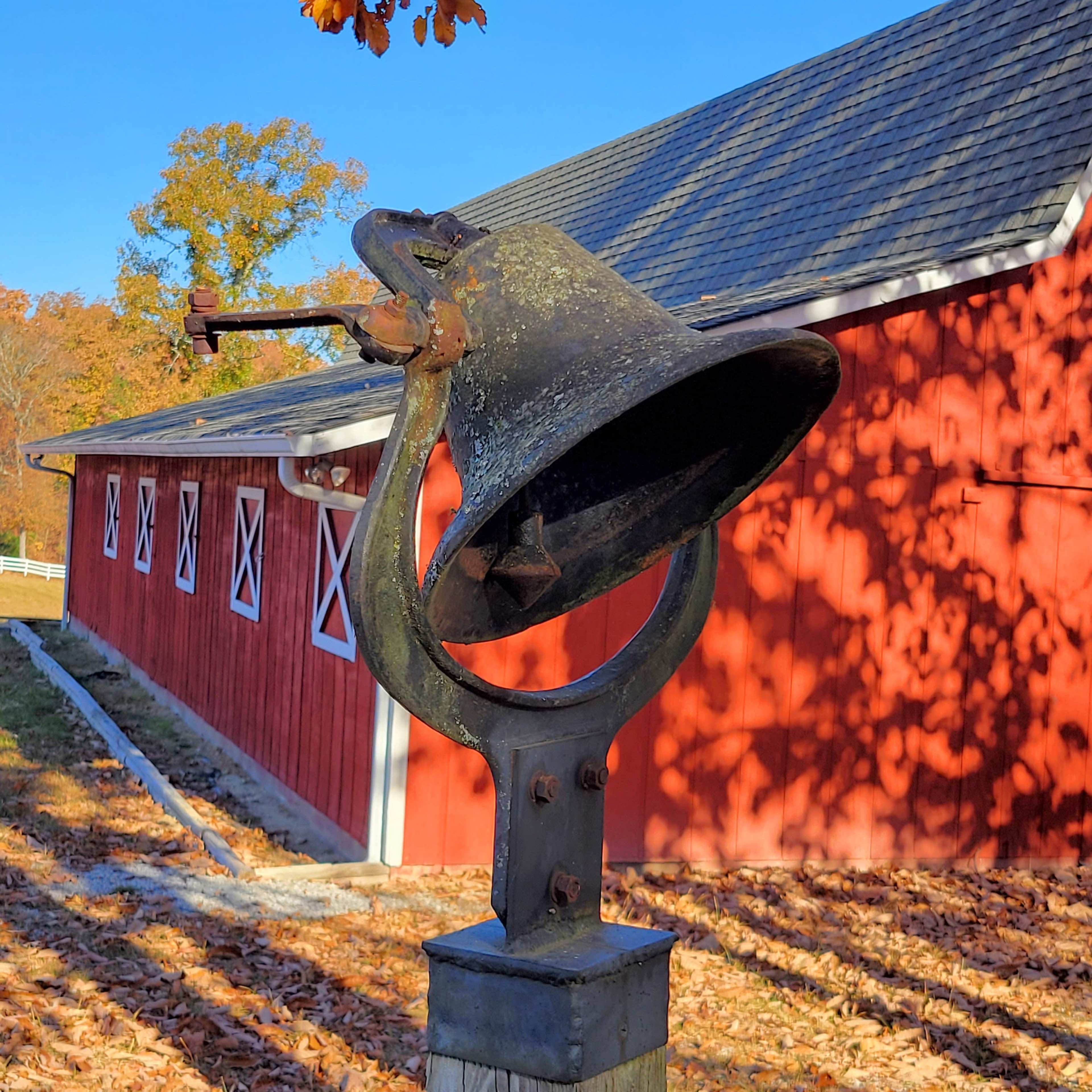 A large, weathered bell is mounted on a post in front of a red barn with white windows.