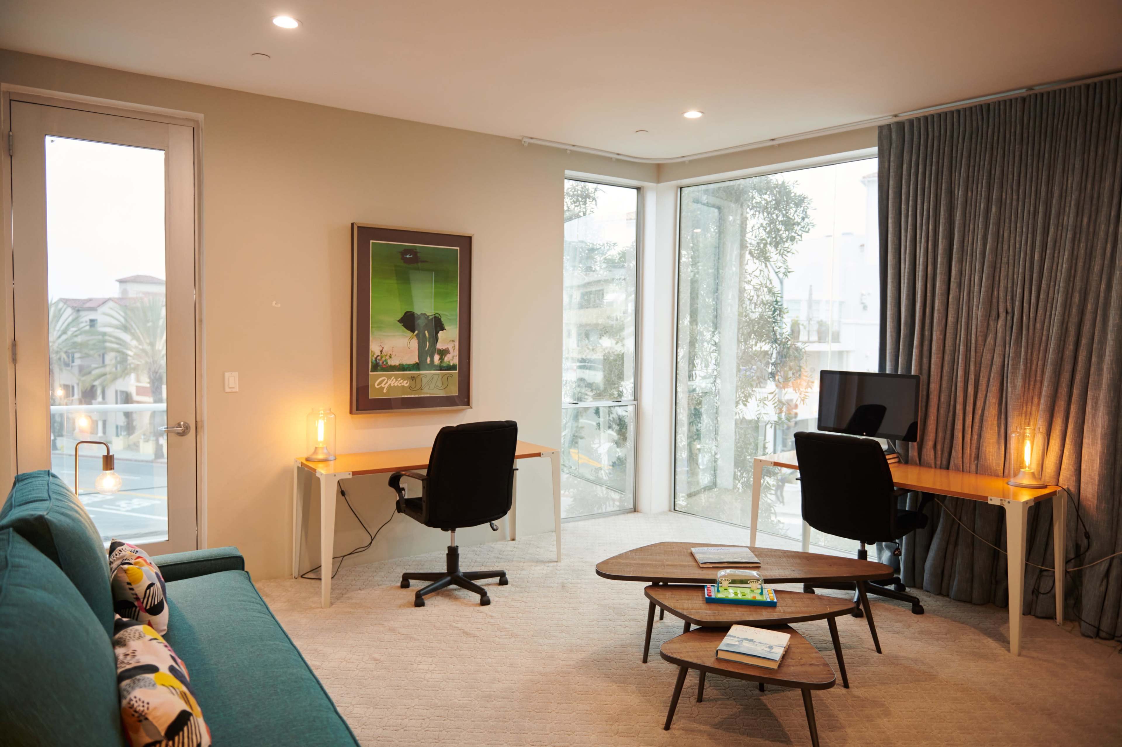 A modern living space featuring a teal sofa, two work desks with chairs, large windows, and a coffee table.