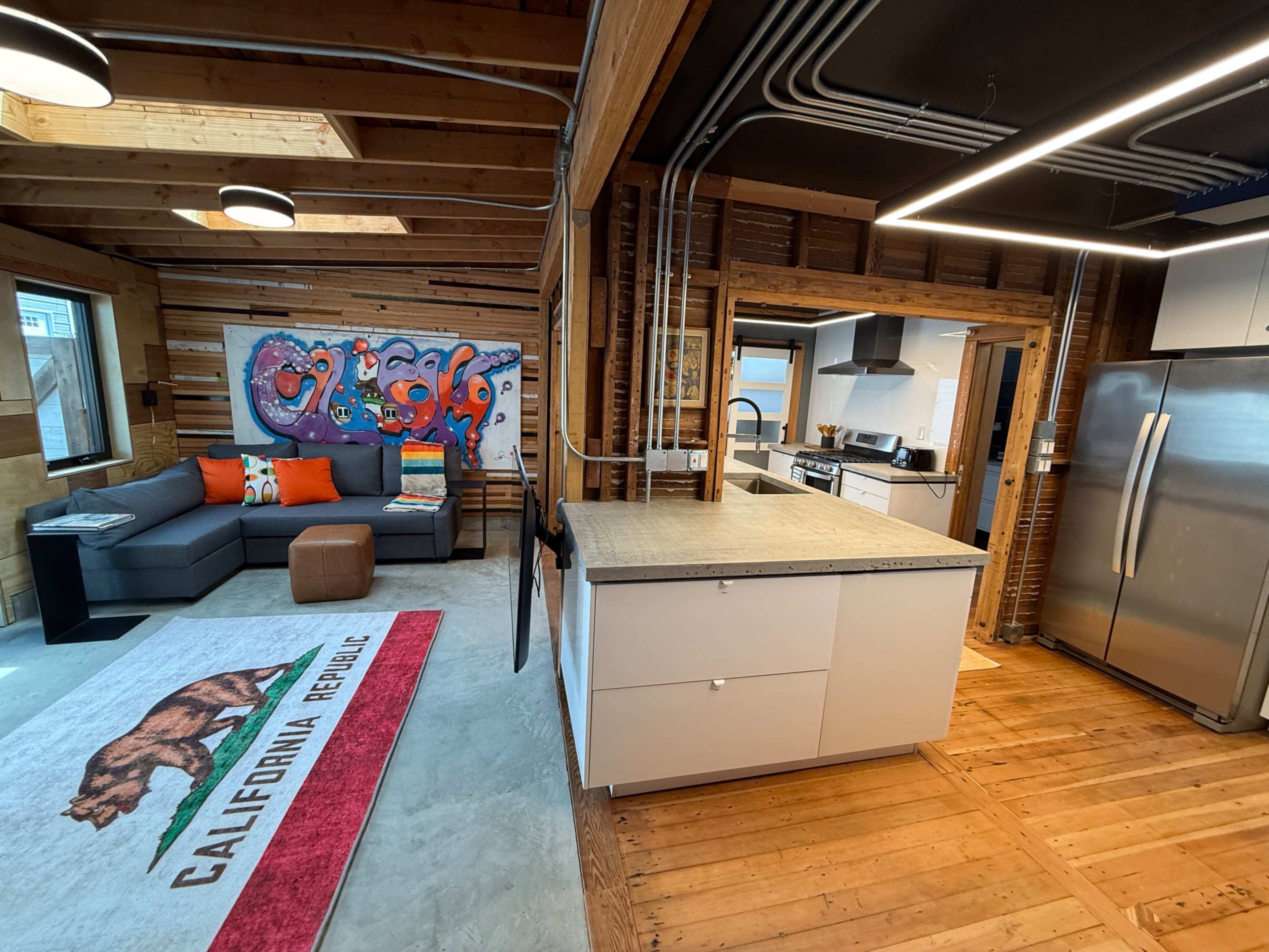 The image shows an open-concept living space featuring a gray sofa, colorful artwork on the wall, and a kitchen with stainless steel appliances and a large island.