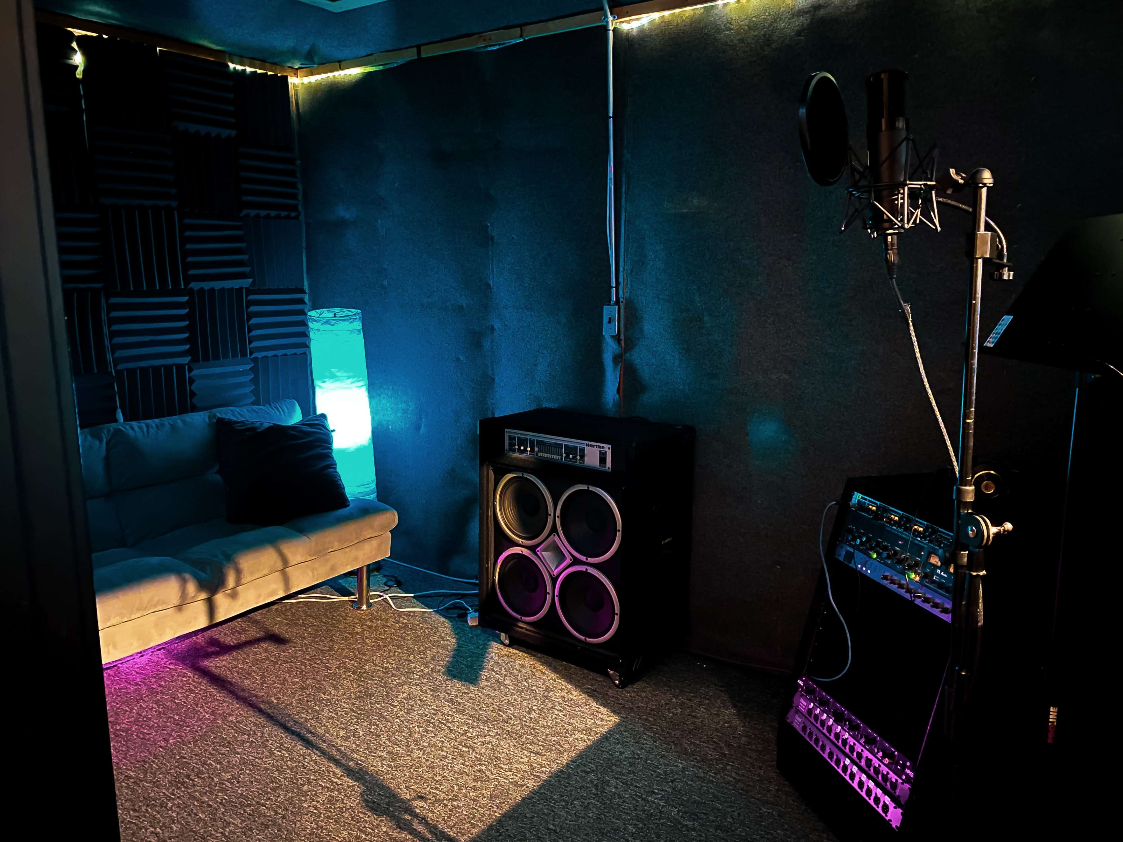 A small soundproof room with a microphone, audio equipment, a couch, and a colorful lamp.
