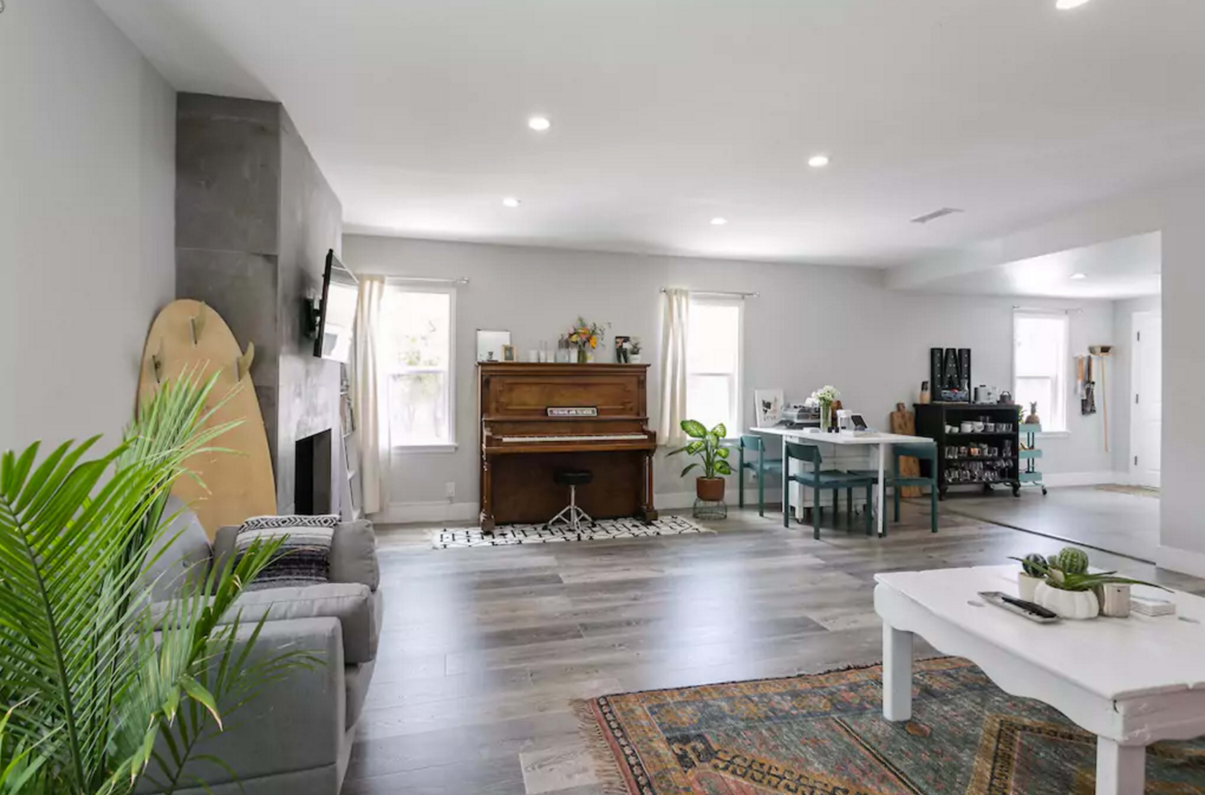 A spacious living area with a piano, a dining table, and a couch, featuring light-colored walls and wooden flooring.