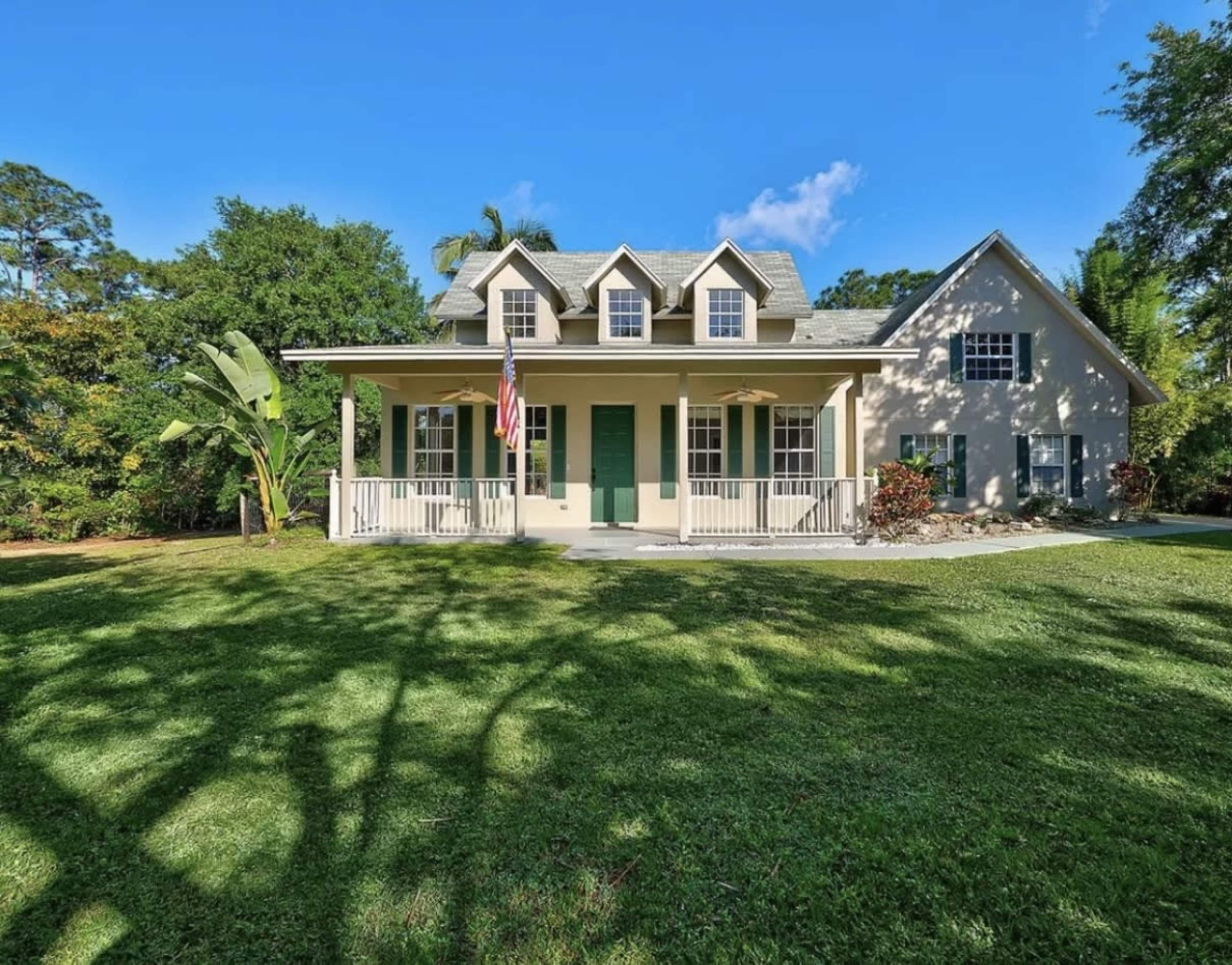 A two-story house with green shutters and an American flag stands amidst a grassy lawn surrounded by trees.