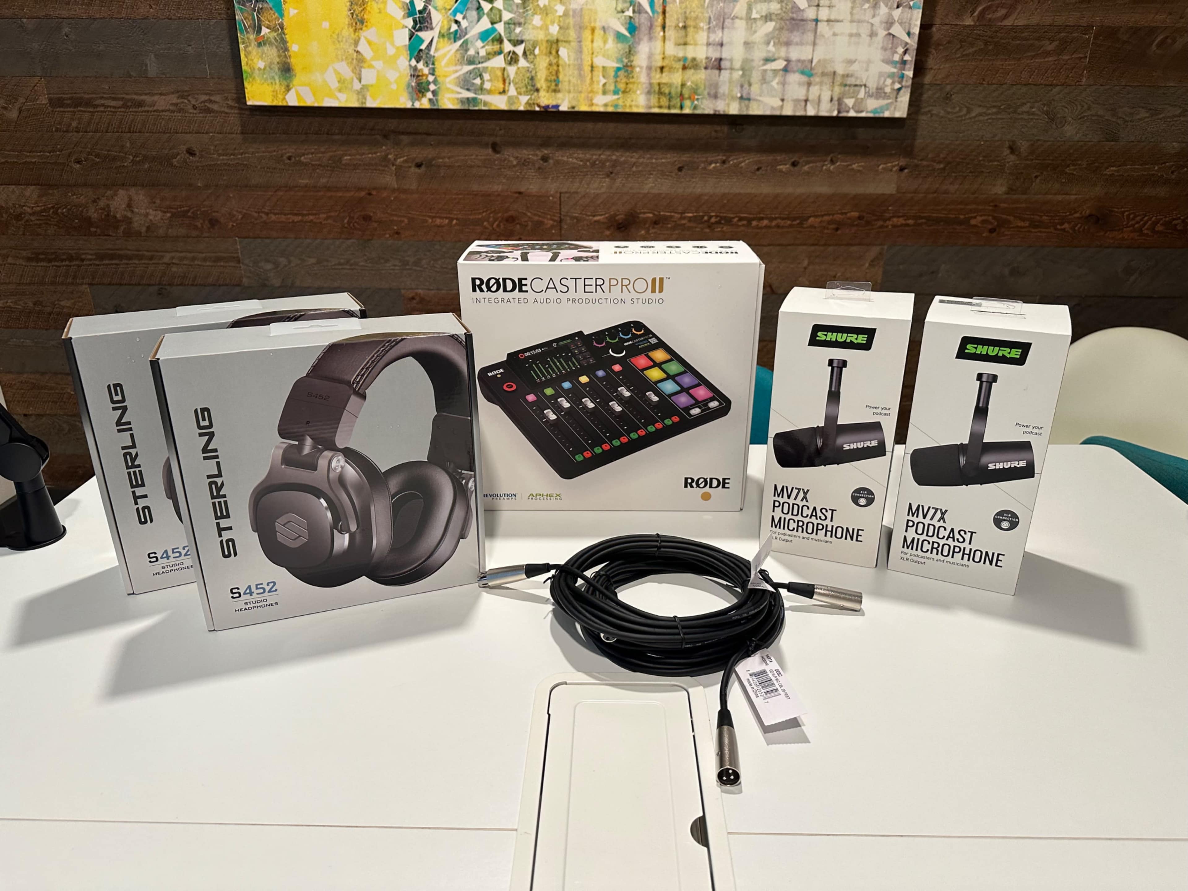 The image shows a collection of audio equipment on a white table, including headphones, a microphone, a podcast mixer, and a cable.