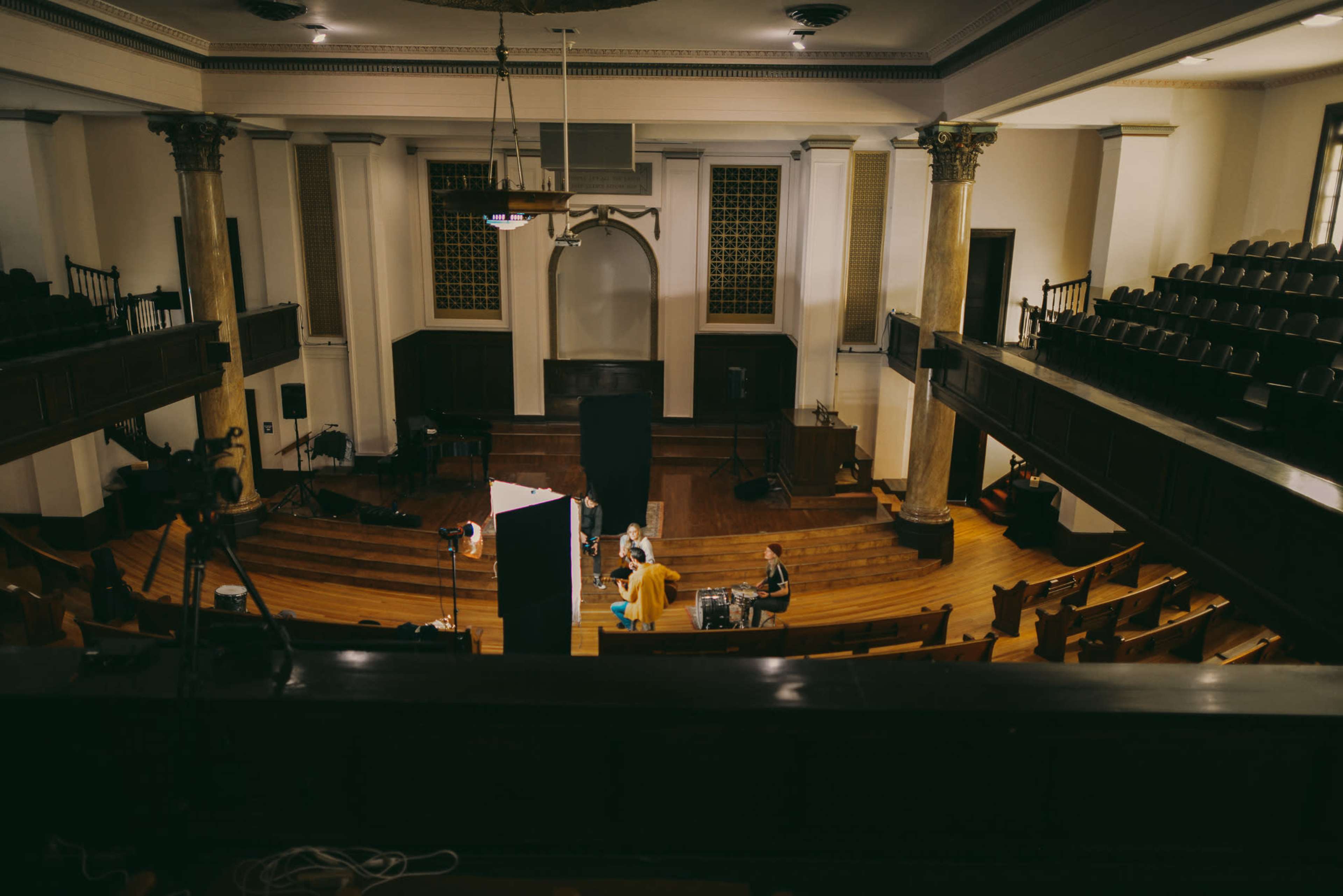 A spacious interior of a theater or hall features wooden seating, tall columns, and crew members setting up for a performance or filming in the center.
