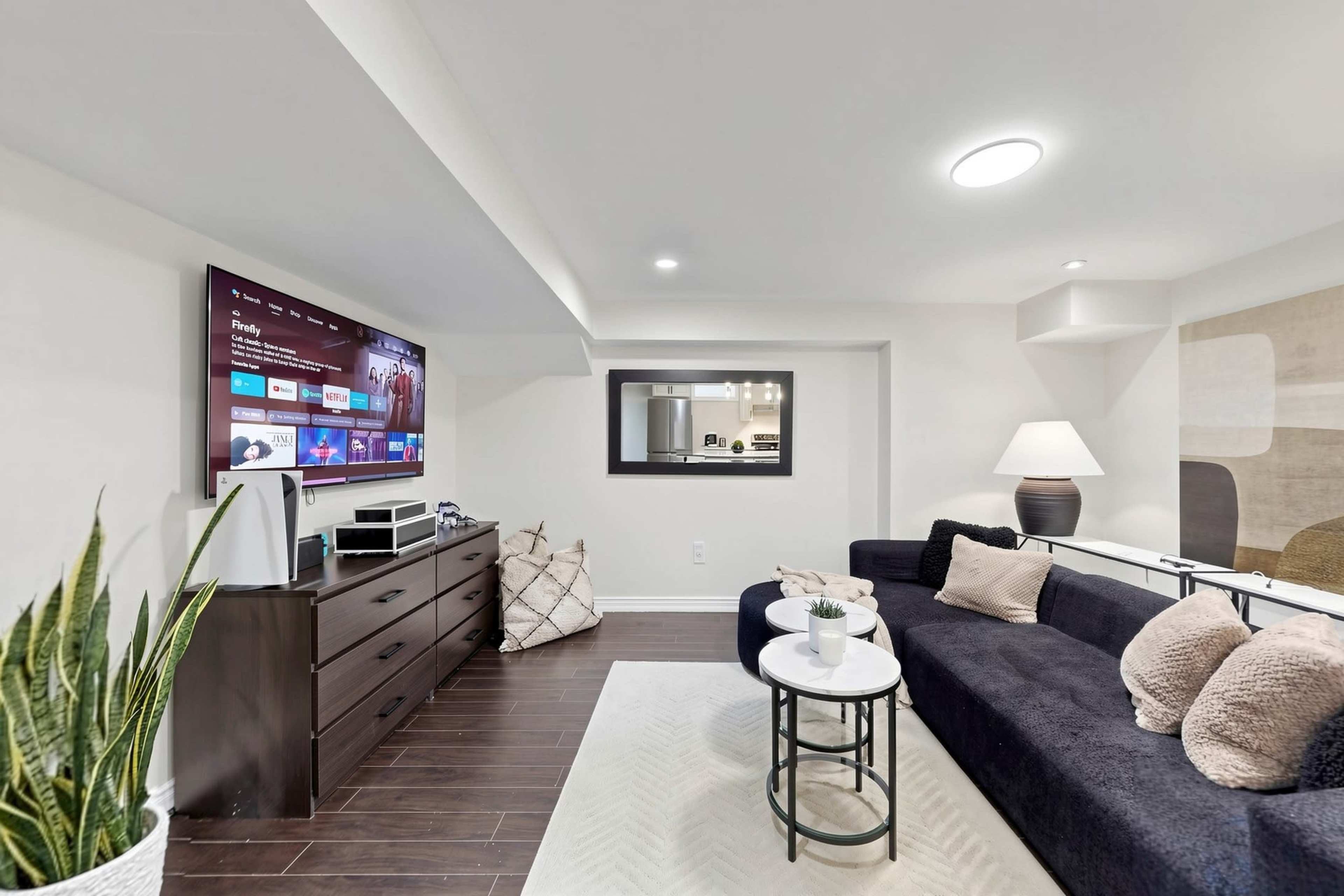 A modern basement living room features a dark sofa, a flat-screen TV on the wall, a coffee table, and decorative plants.