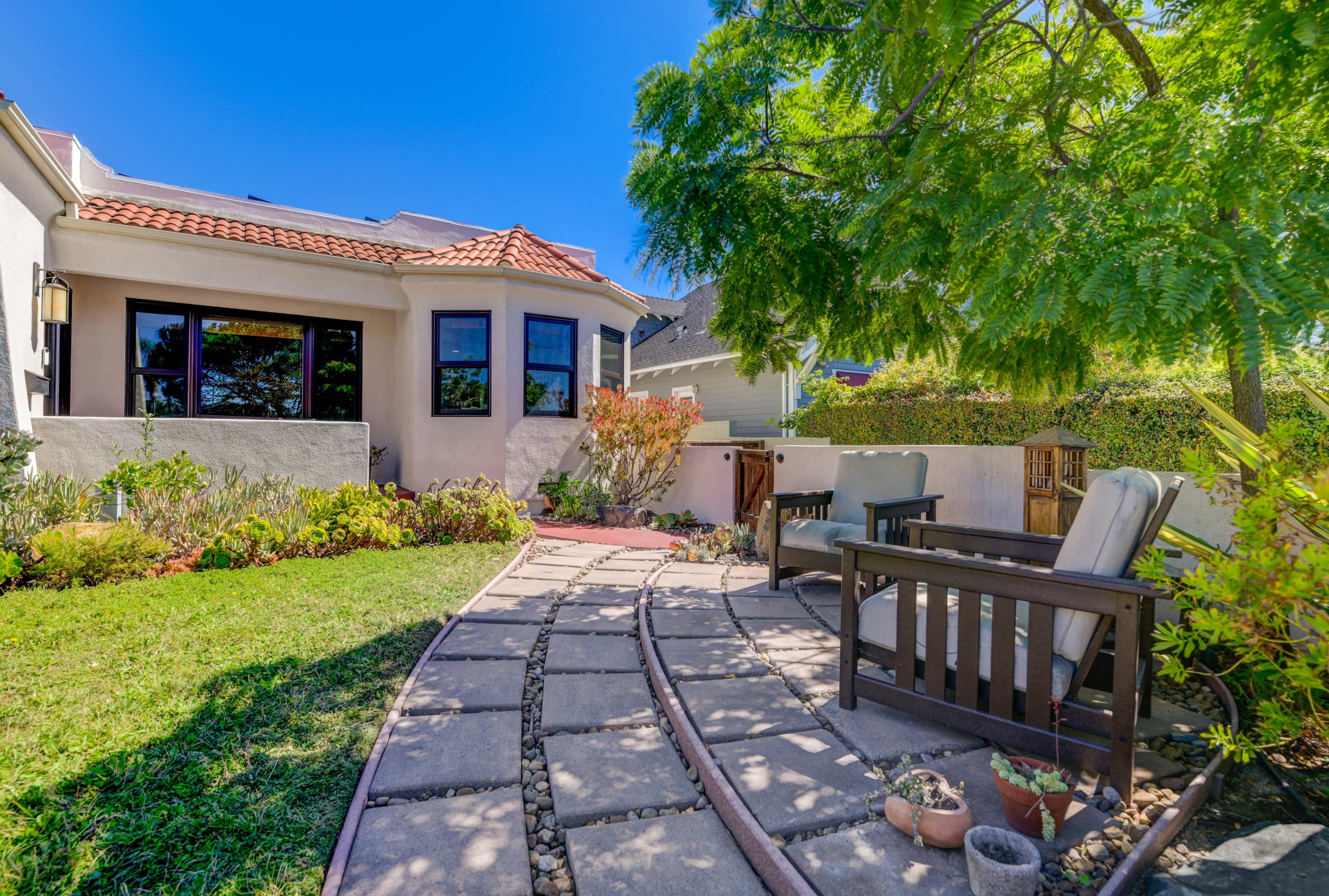 A landscaped backyard with a stone pathway leading to a seating area and a hot tub surrounded by greenery.