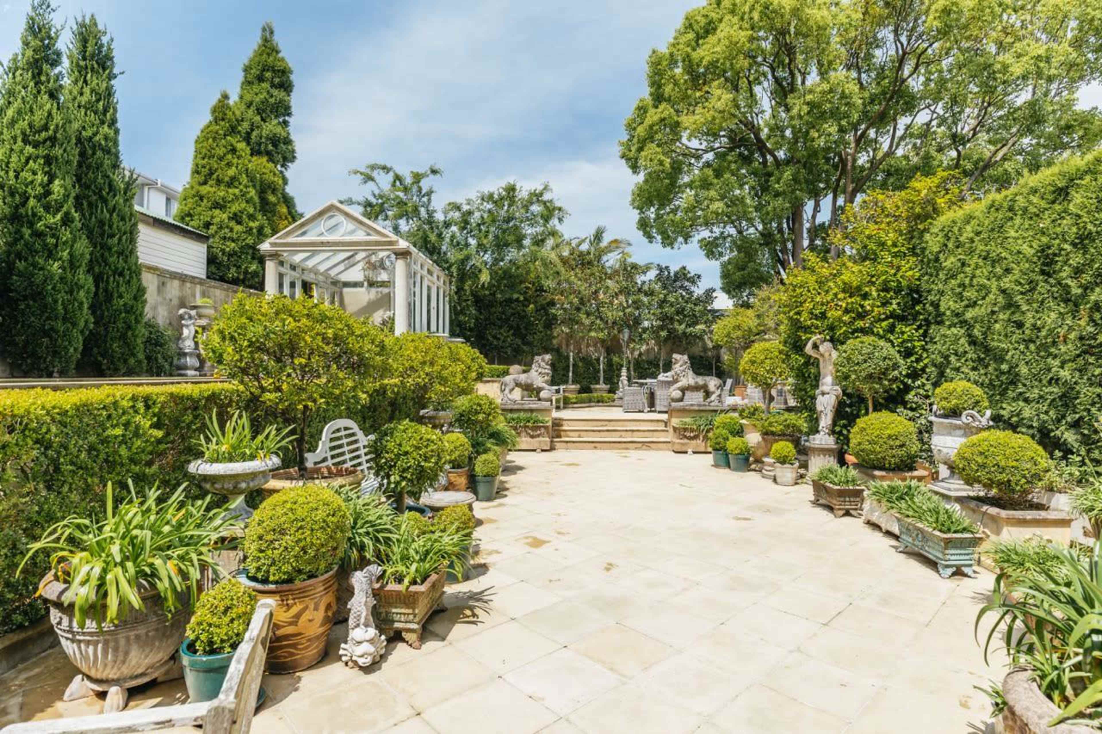 A landscaped outdoor garden area with stone pathways, manicured hedges, and potted plants arranged around ornamental sculptures.