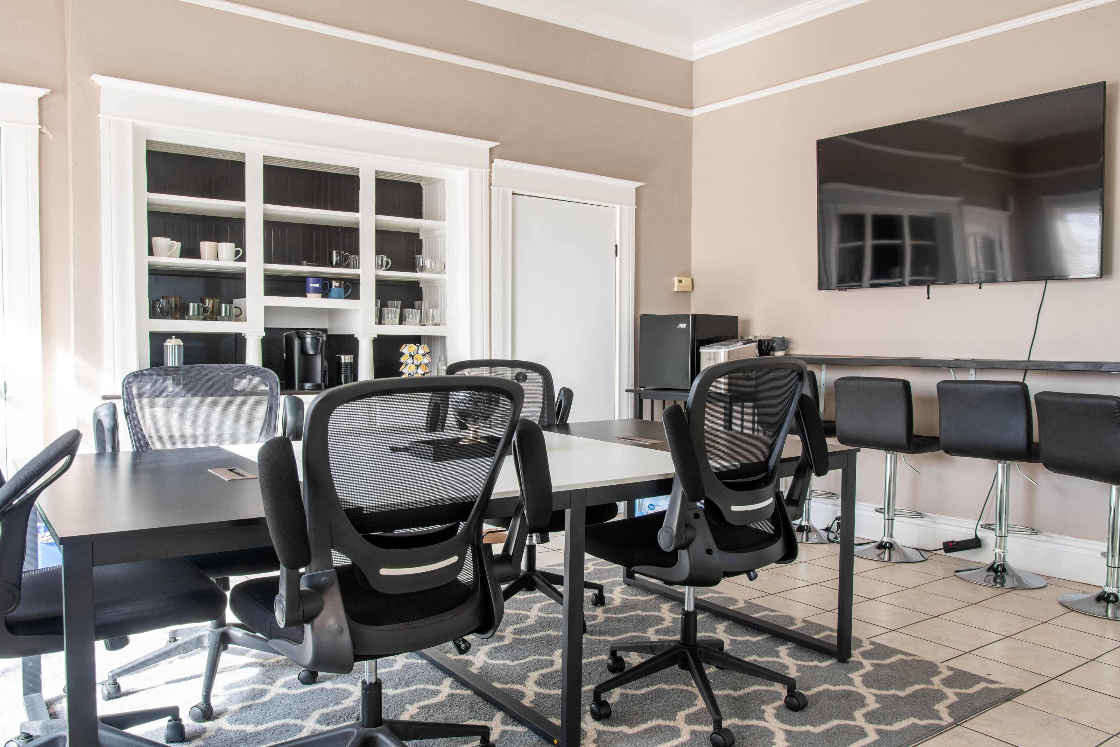 A conference room features a large table surrounded by several black chairs, with a wall-mounted screen and a storage cabinet in the background.