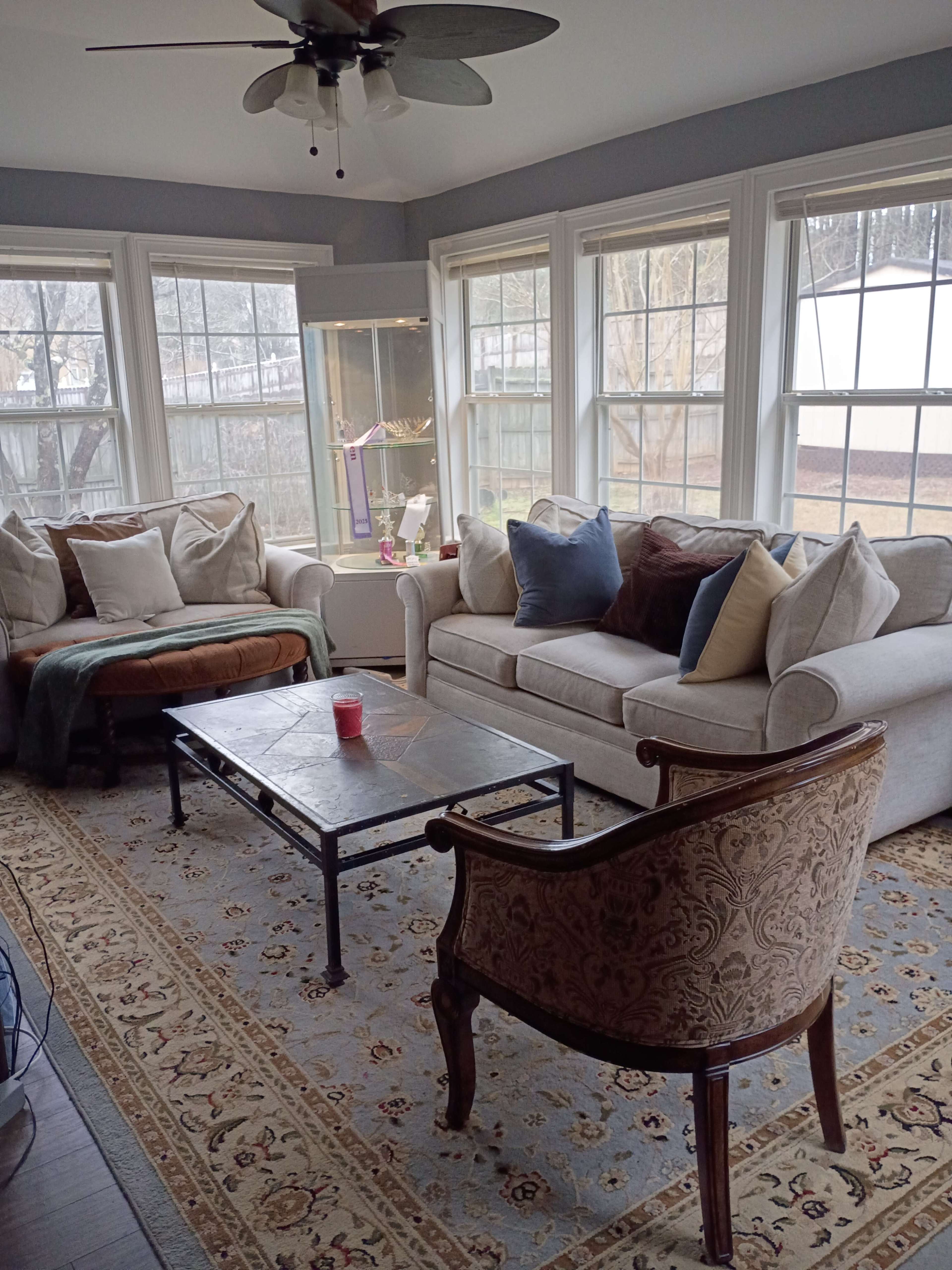 The image shows a sunroom with a light-colored couch, an armchair, and large windows that illuminate the space.