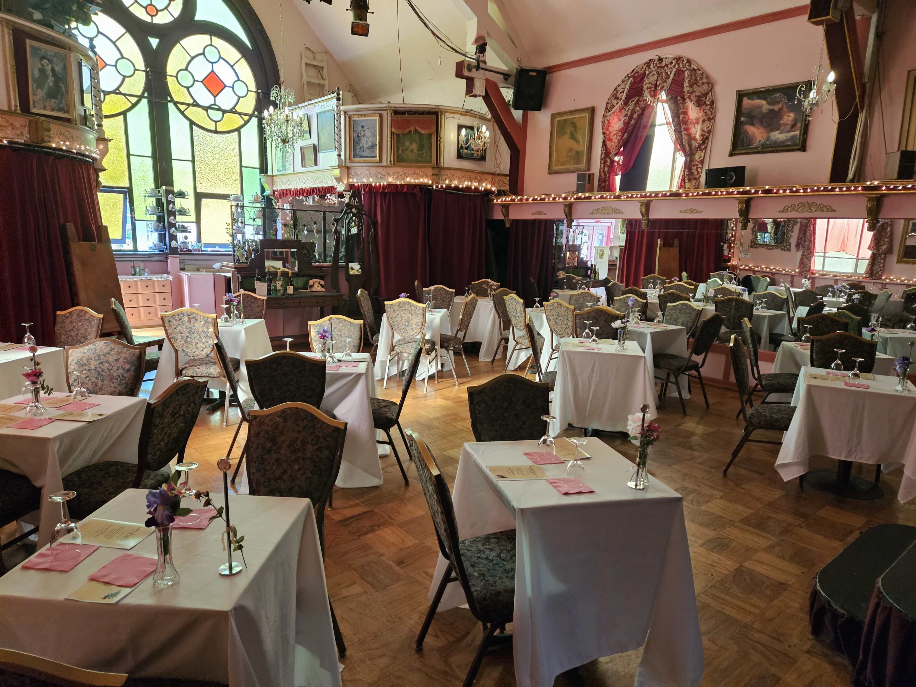 A dining area with tables covered in white tablecloths, decorated with small vases of flowers, and surrounded by framed artwork and stained glass windows.
