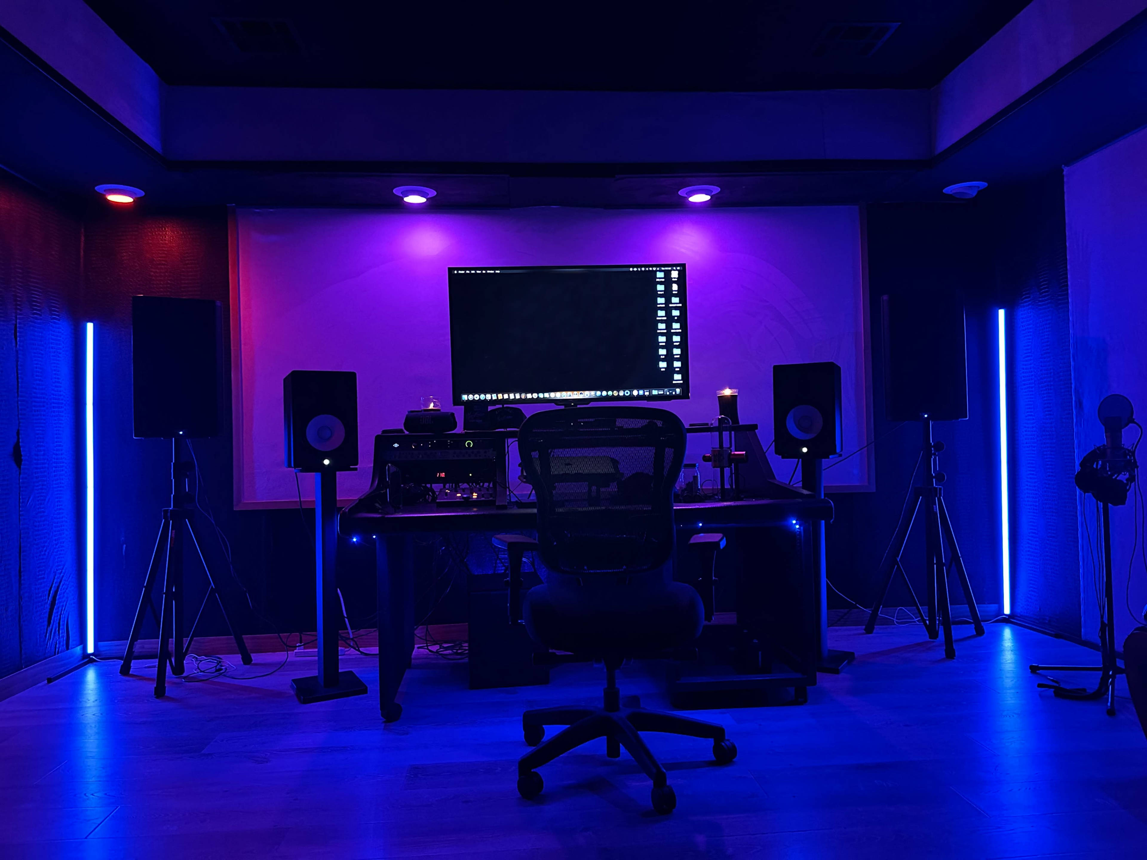 A dimly lit music studio features a central desk with a computer monitor, surrounded by speakers and illuminated by colorful LED lights.