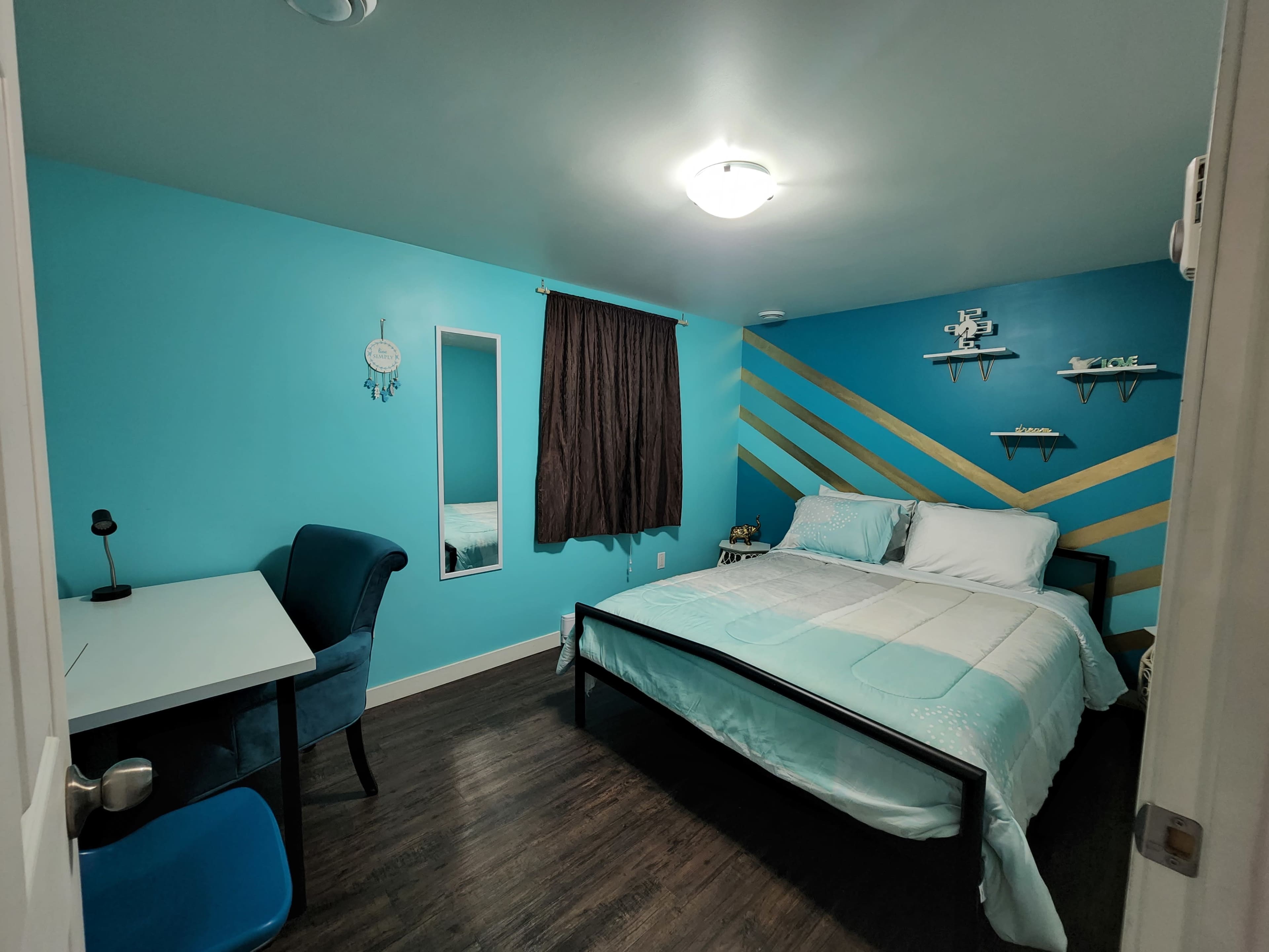 The image shows a bedroom with a large bed, a desk with a chair, and teal walls featuring decorative shelves.