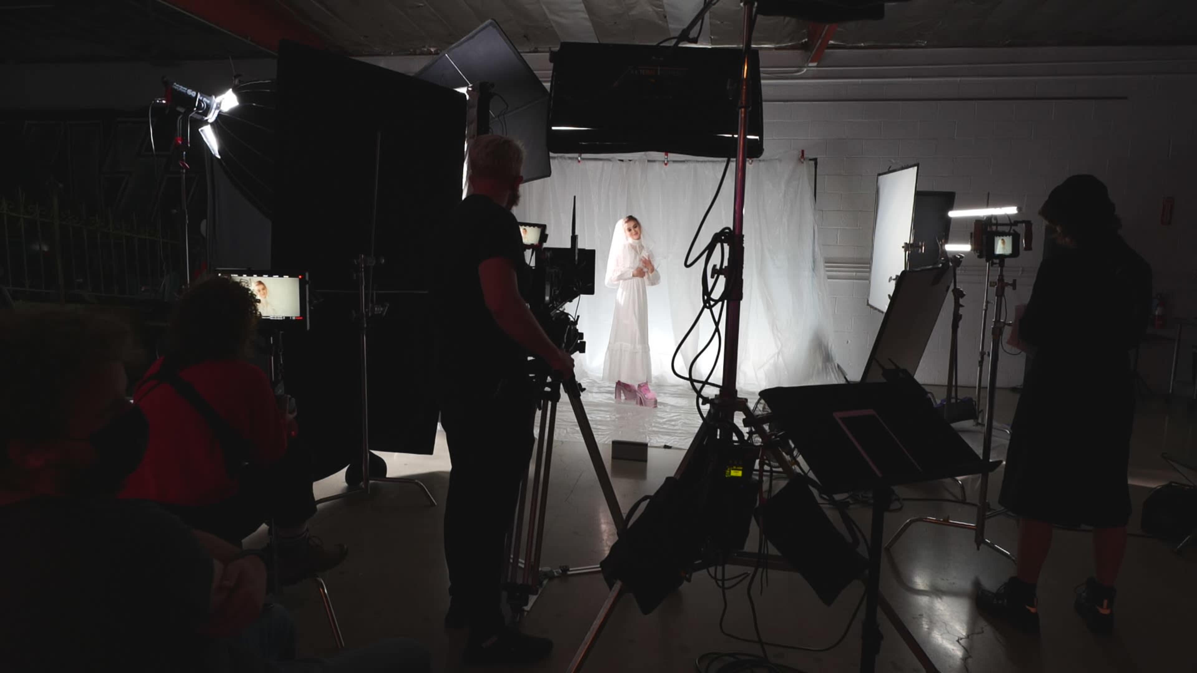 A film crew sets up equipment in a studio to shoot a model wearing a white outfit against a backdrop of white fabric.