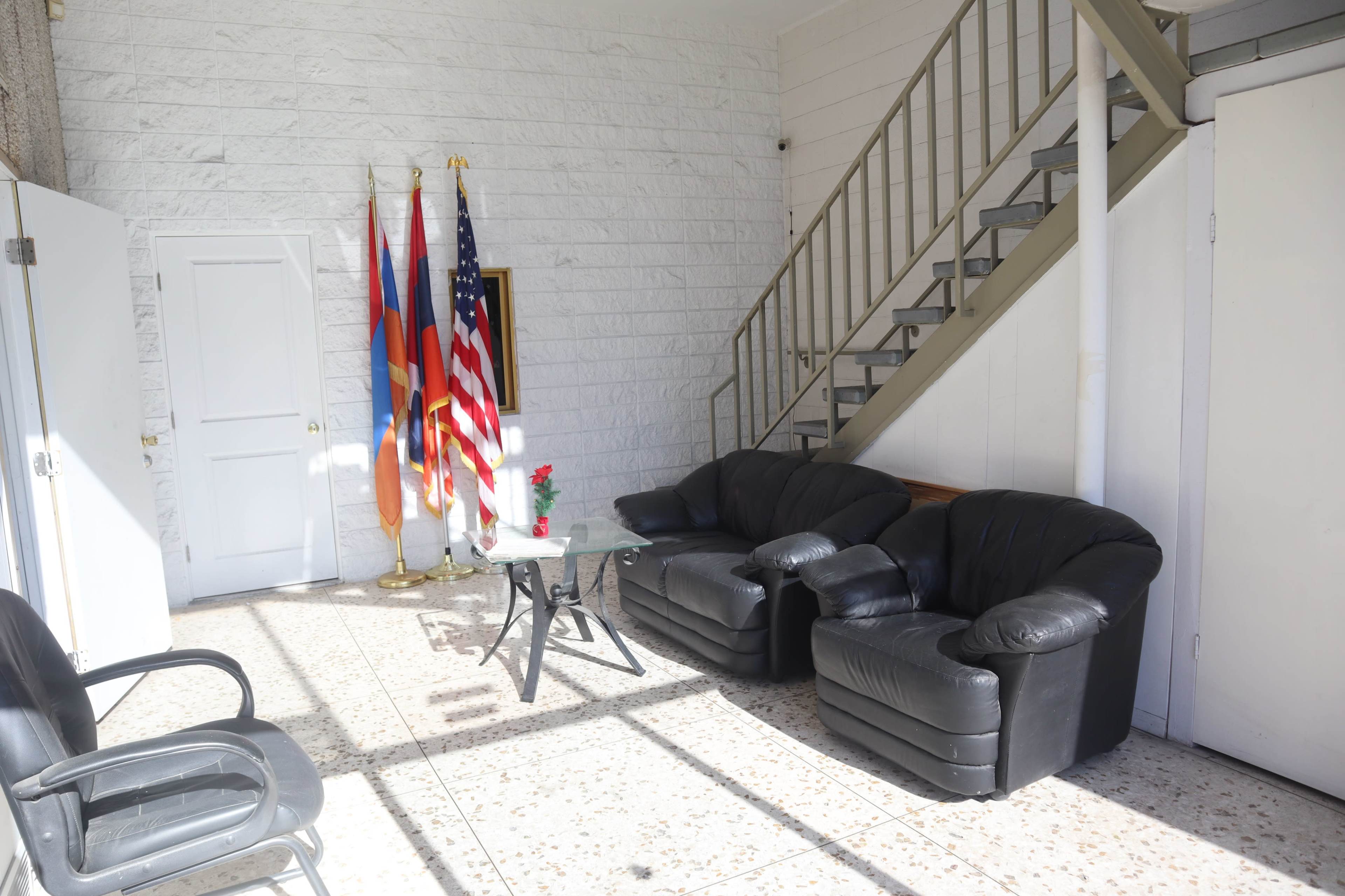 A well-lit foyer with two black armchairs, a glass table, and three flags displayed on stands, alongside a staircase leading to an upper level.