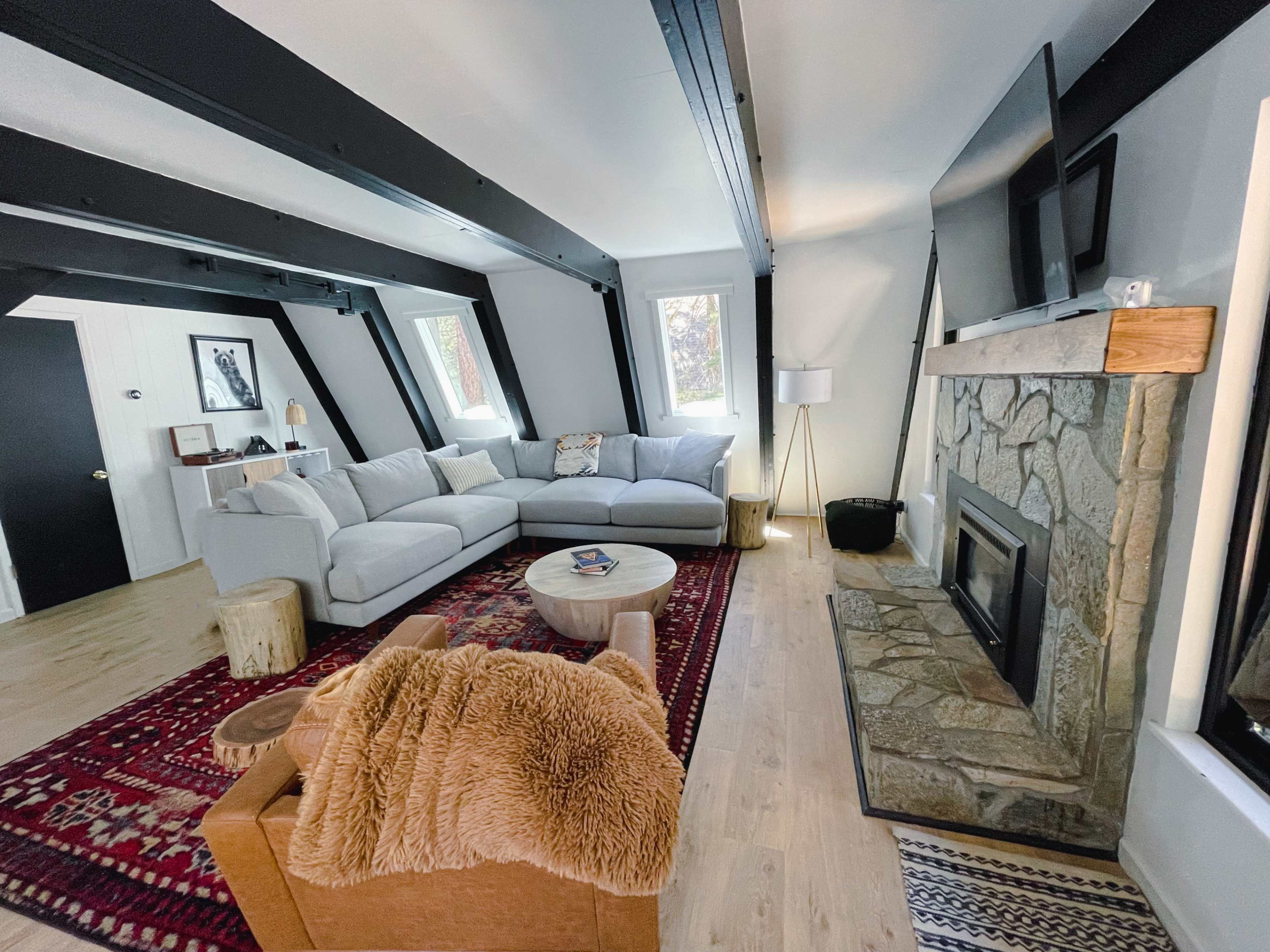 A modern living room featuring a large sectional sofa, a stone fireplace, a round coffee table, and a plush armchair, all set against a backdrop of exposed beams and hardwood flooring.