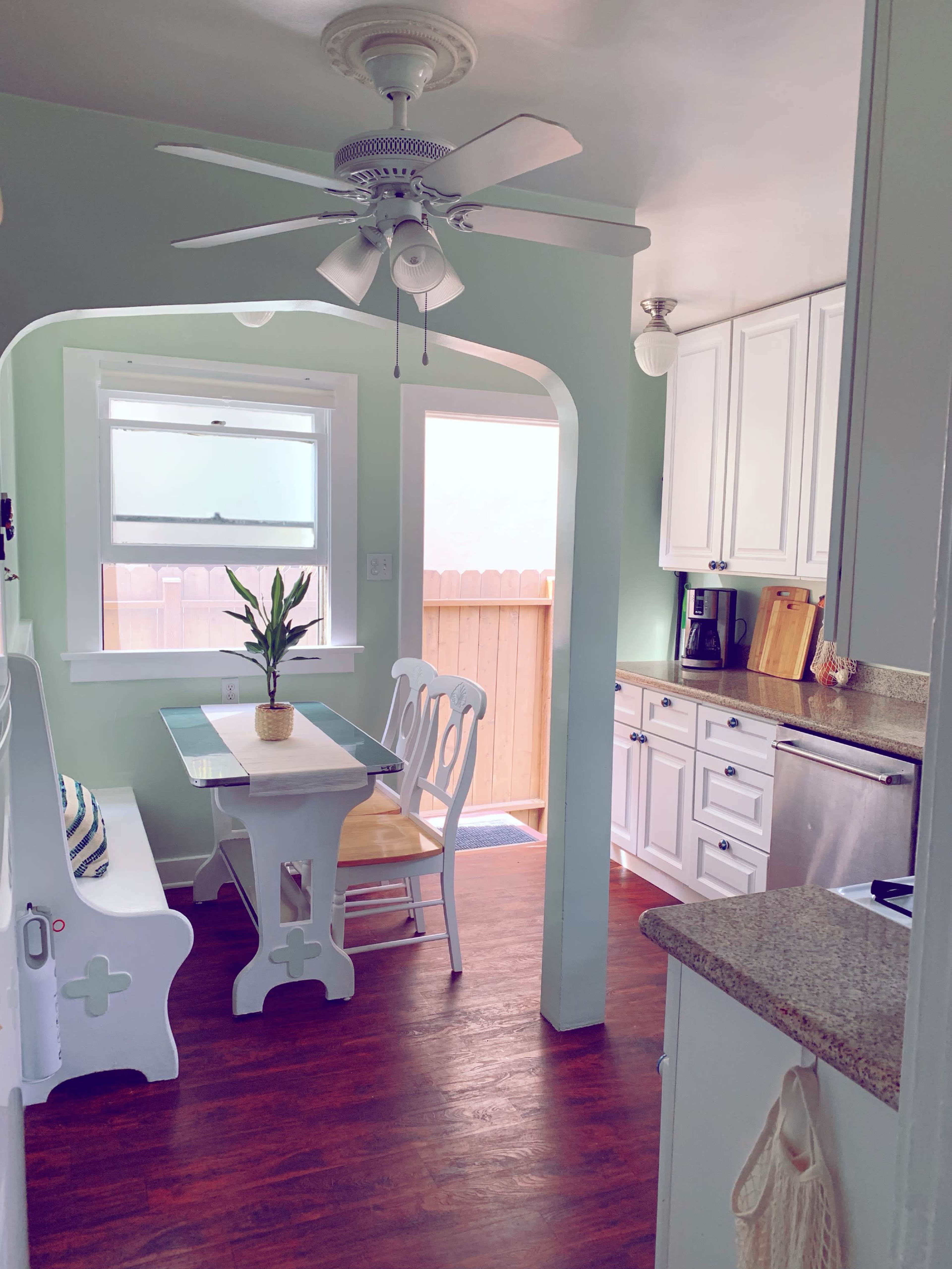 The image shows a compact kitchen area with a small table and chairs, a ceiling fan, and pale green walls, leading to an outdoor entrance.