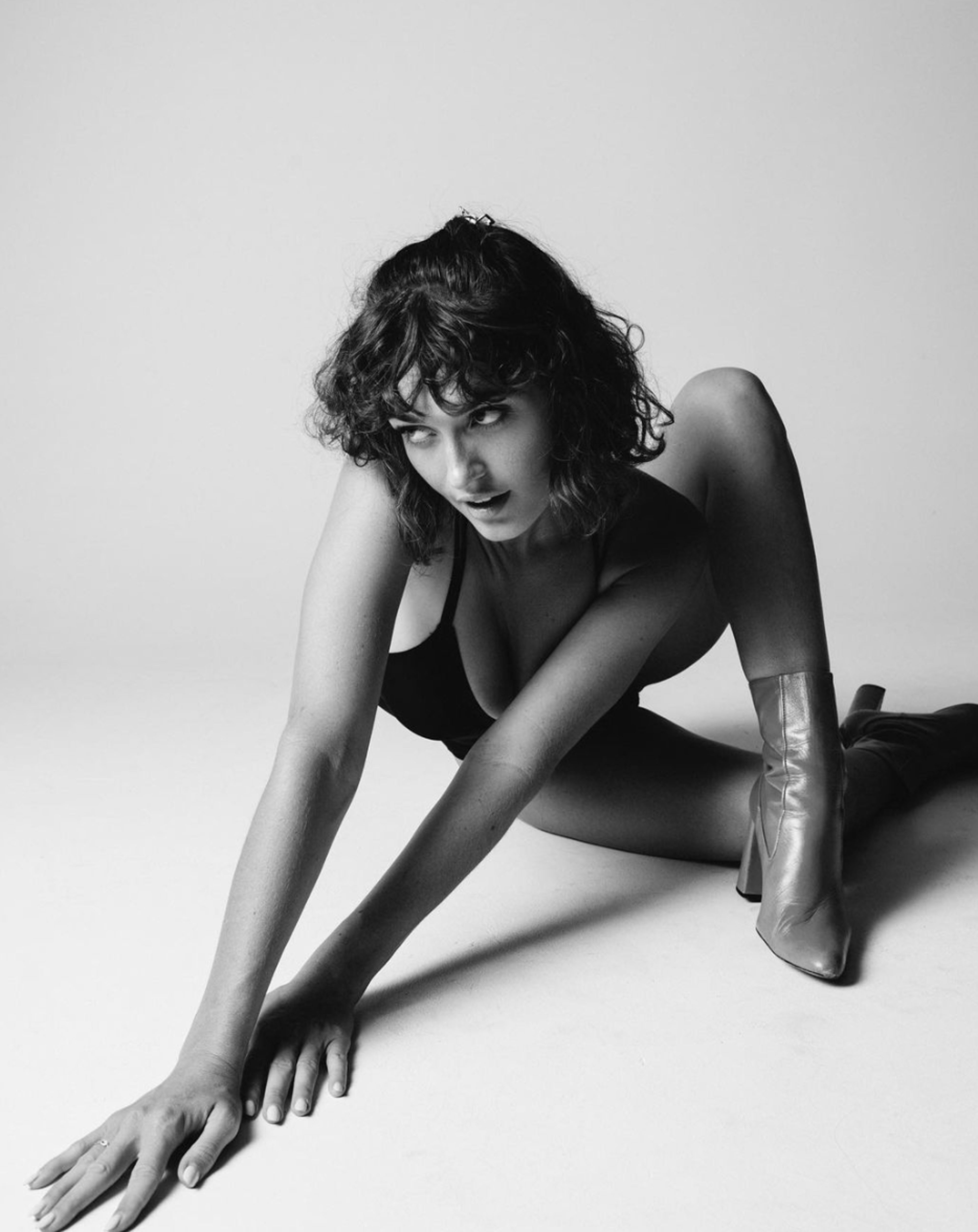 A person with curly hair poses on the floor in a black outfit and high-heeled boots.