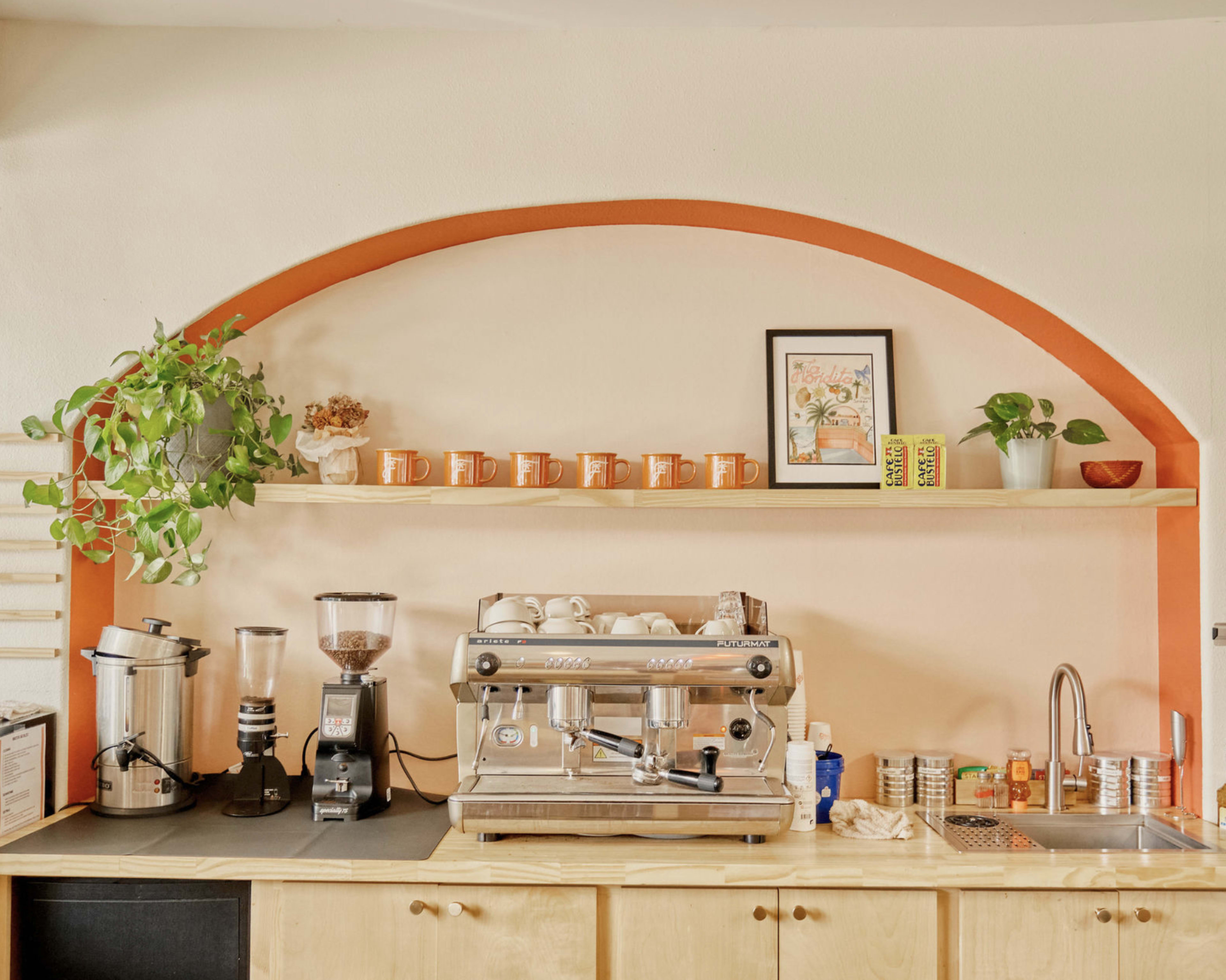 A coffee station features an espresso machine, a grinder, and various mugs displayed on a shelf against a pastel wall.