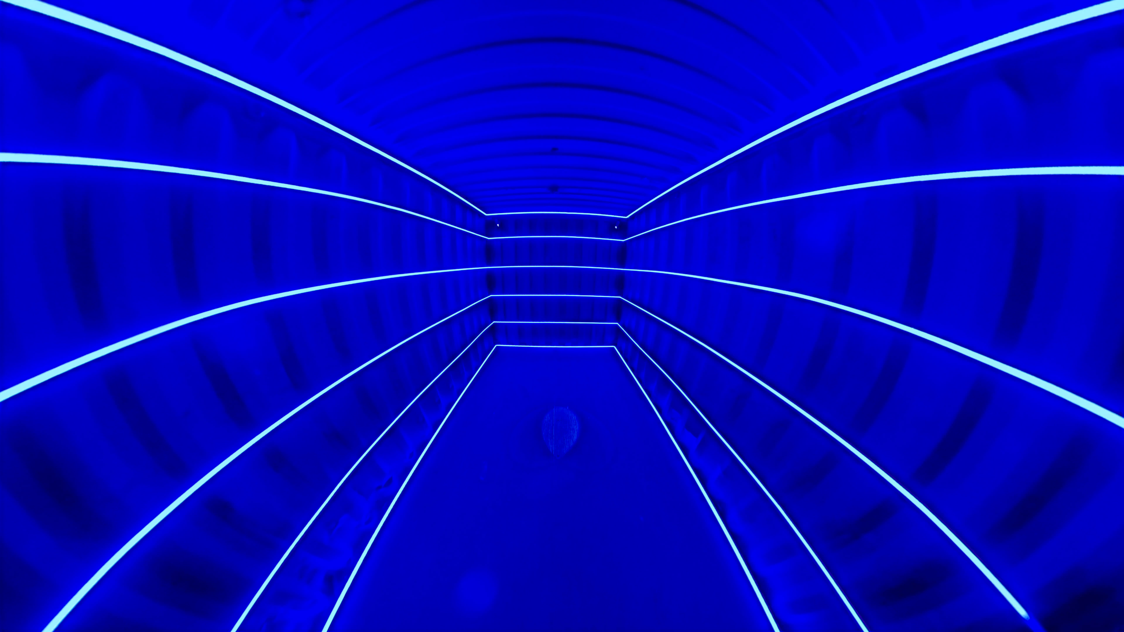 The image shows a tunnel with curved walls illuminated by blue lights and white lines creating a geometric pattern.