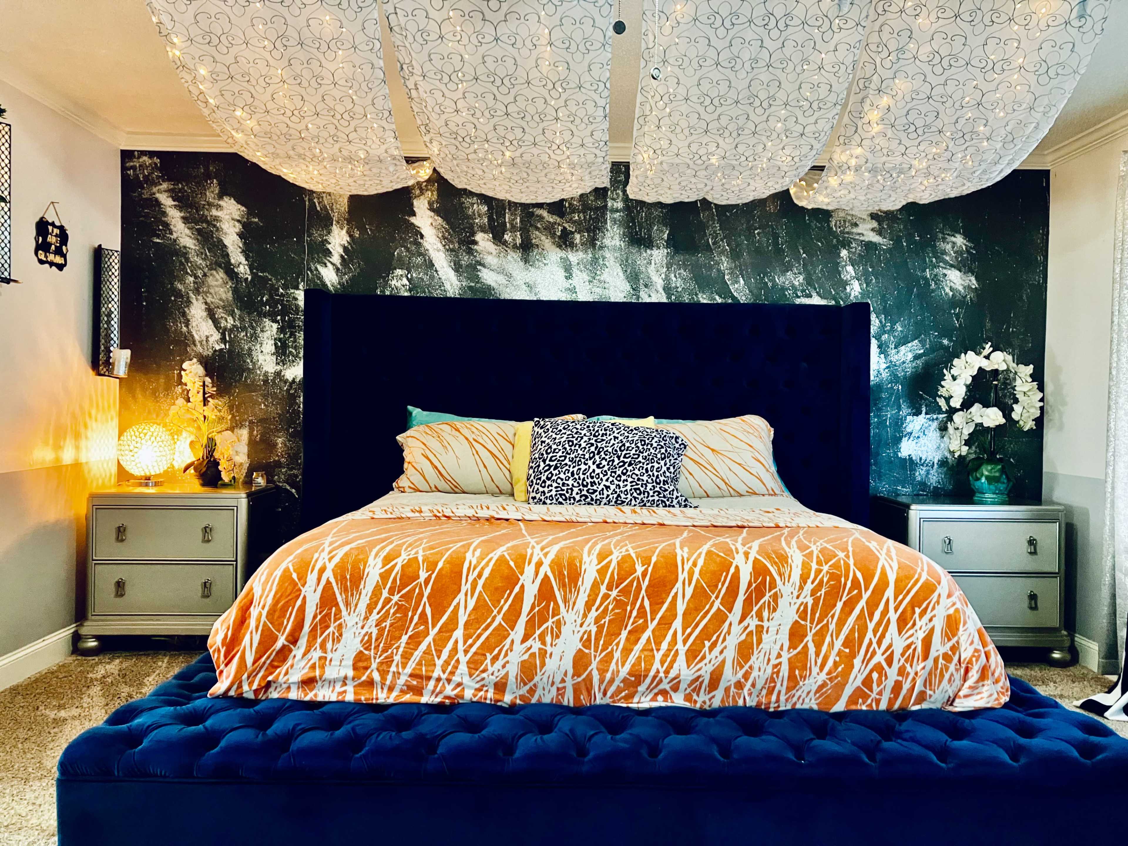 The image shows a well-decorated bedroom featuring a large blue bed with an orange and white patterned comforter, against a textured black wall, with decorative lights and floral arrangements on bedside tables.