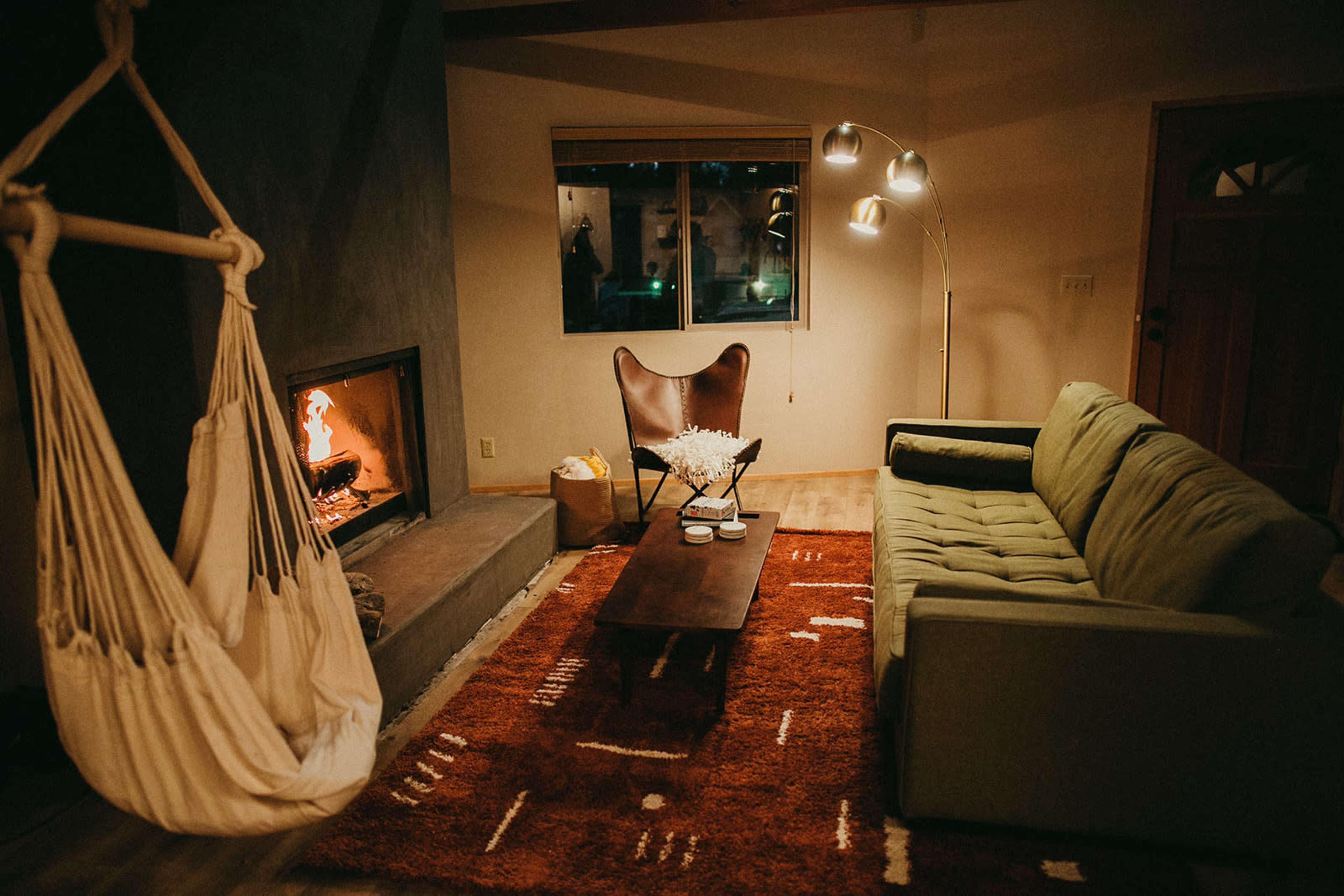The living room features a green sofa, a wooden coffee table, a hanging chair, and a fireplace, all set against a backdrop of warm lighting and a rustic rug.
