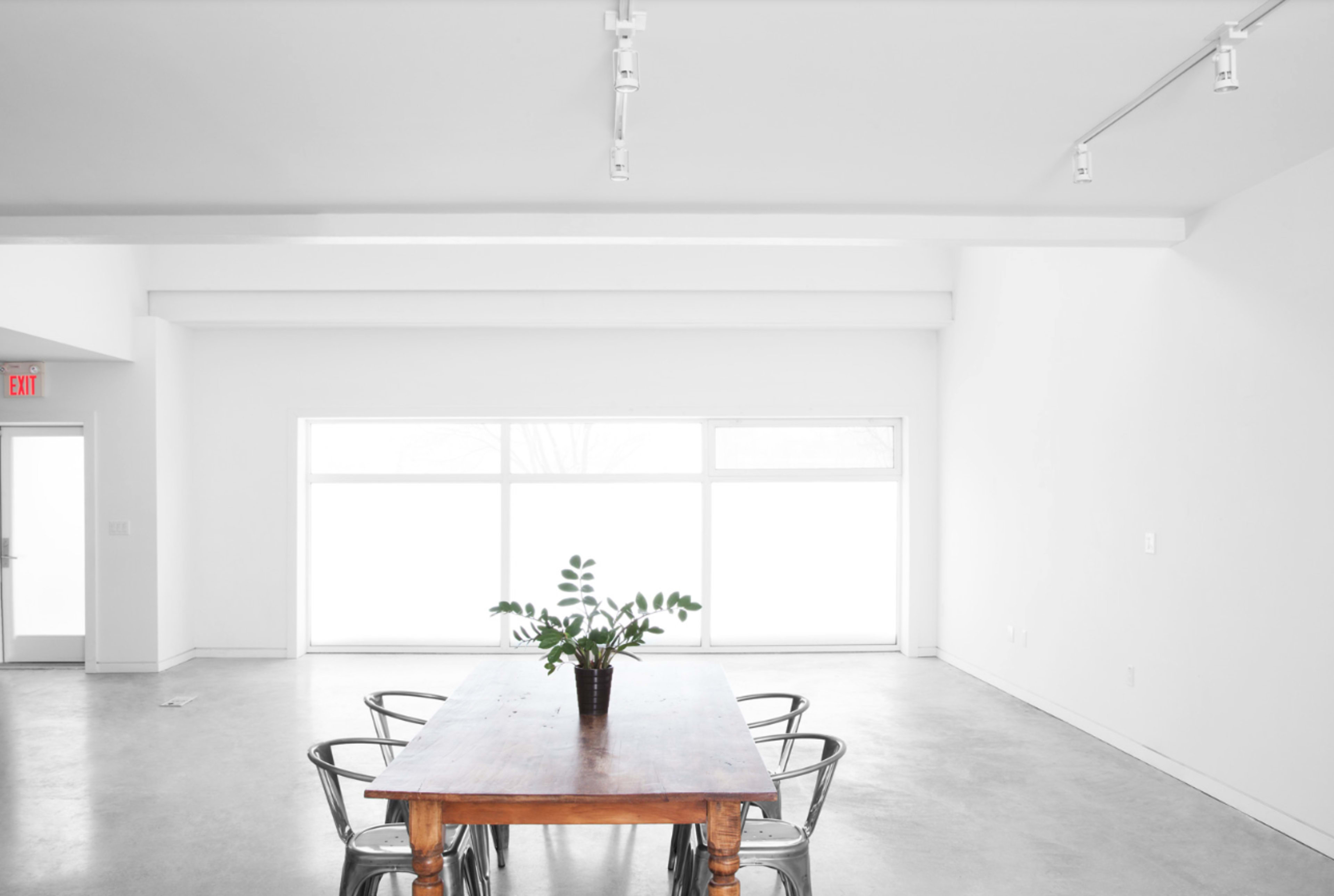 A wooden dining table with metal chairs sits in a bright, empty room featuring large windows.