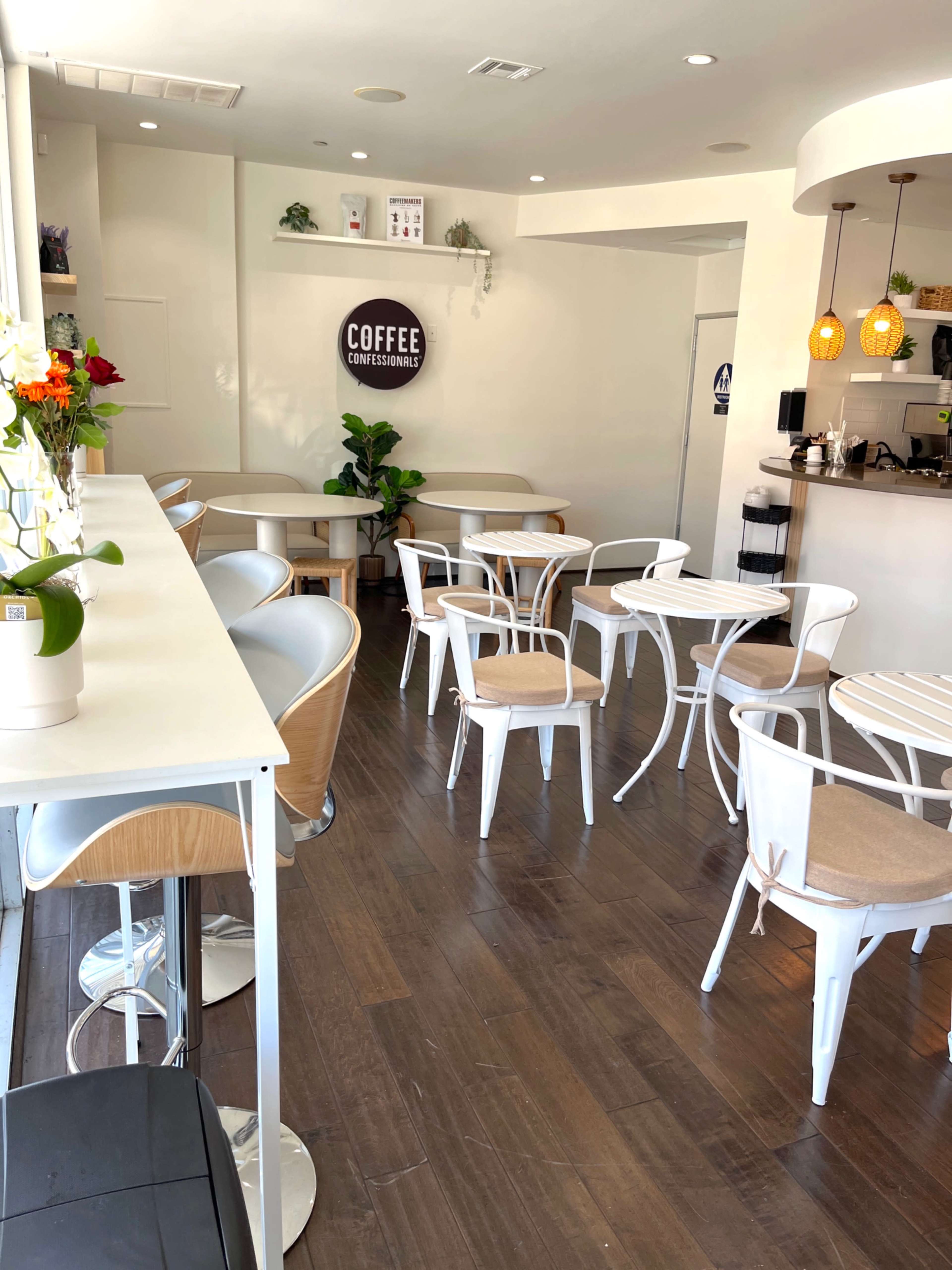 The image shows a modern coffee shop interior with white and wooden furniture, including tables and chairs, and a counter area in the background.