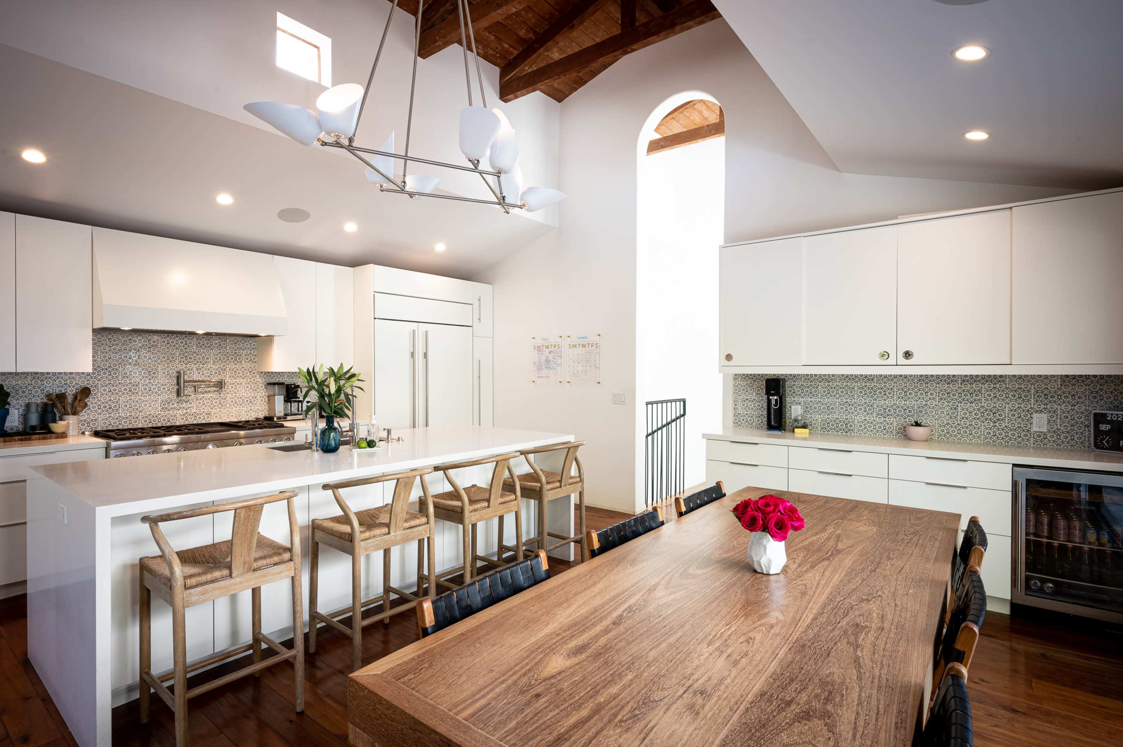 A spacious kitchen features a large island with bar stools, white cabinetry, and a dining table adorned with a vase of flowers.