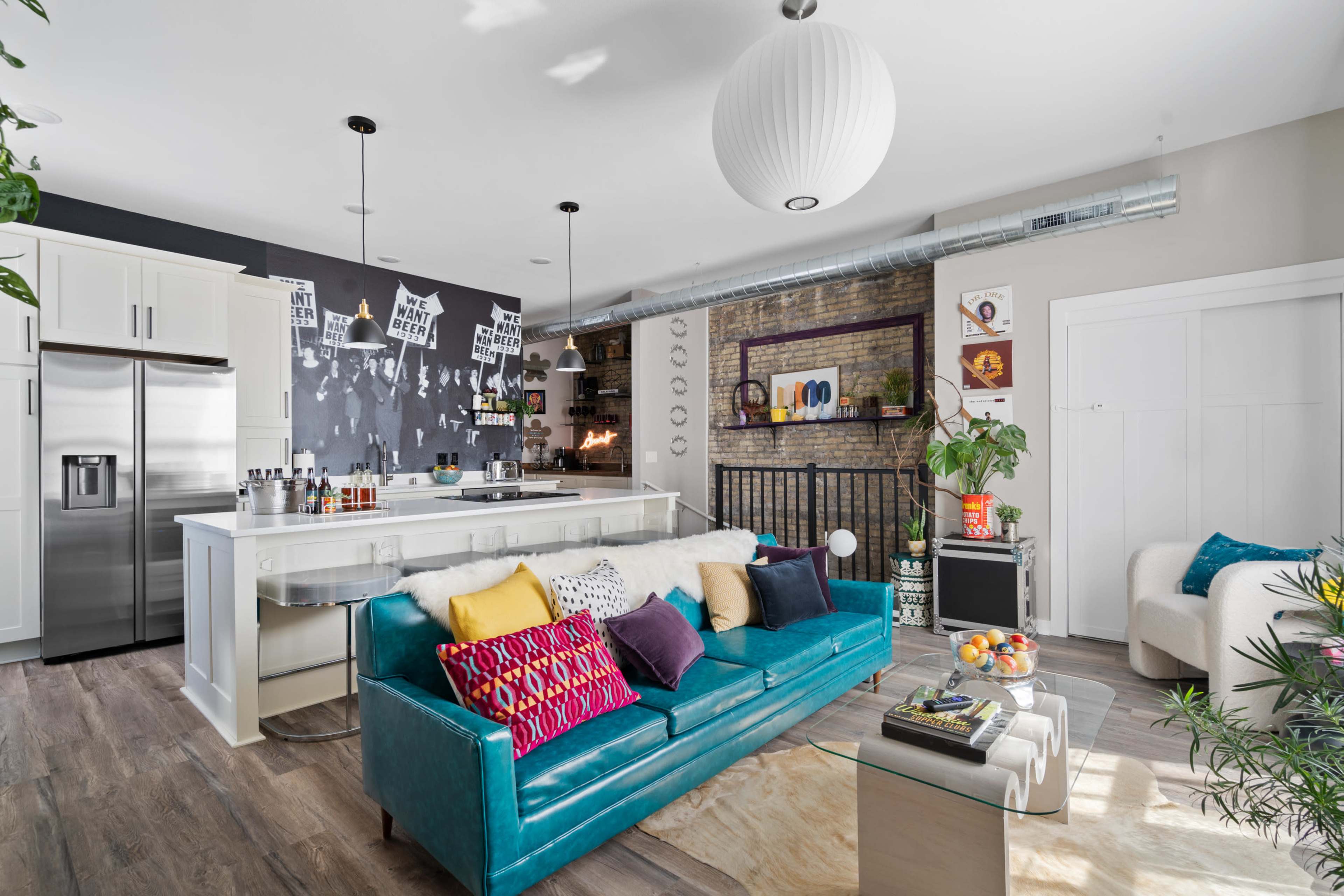 A modern living room features a teal couch, a white kitchen island, an exposed brick wall with decor, and various decorative elements throughout the space.