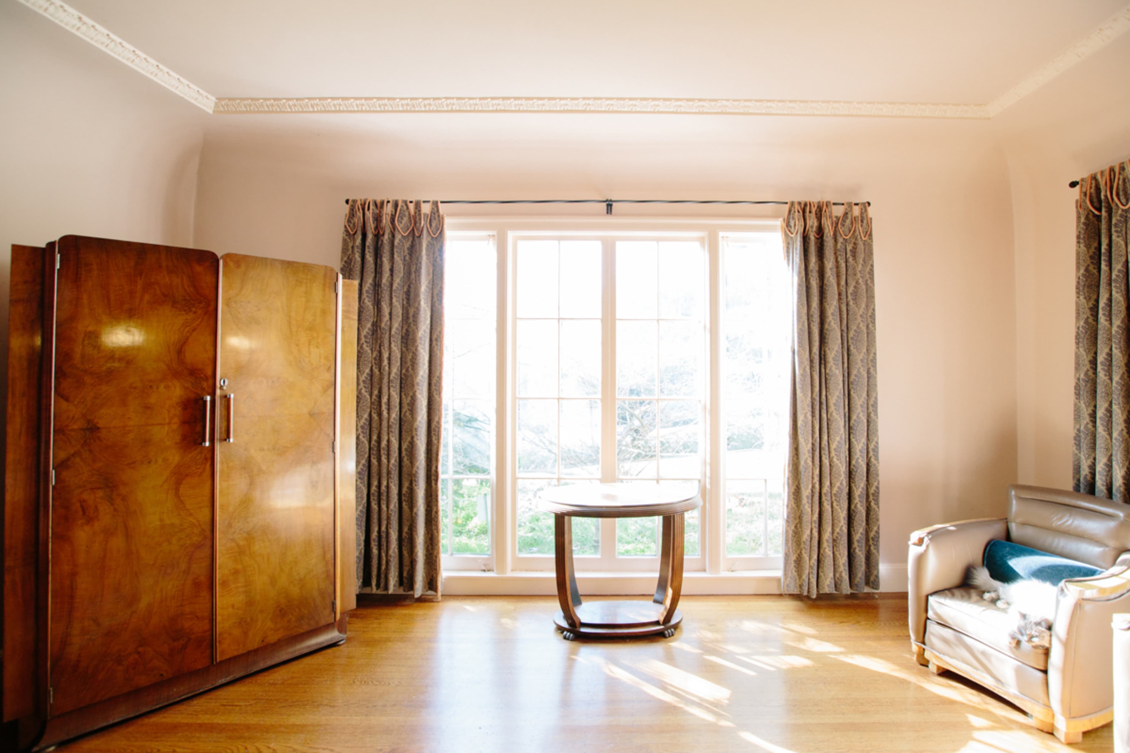 The image shows a room with wooden furniture, including a large wardrobe, a small round table, and a couch, illuminated by light coming through large windows.