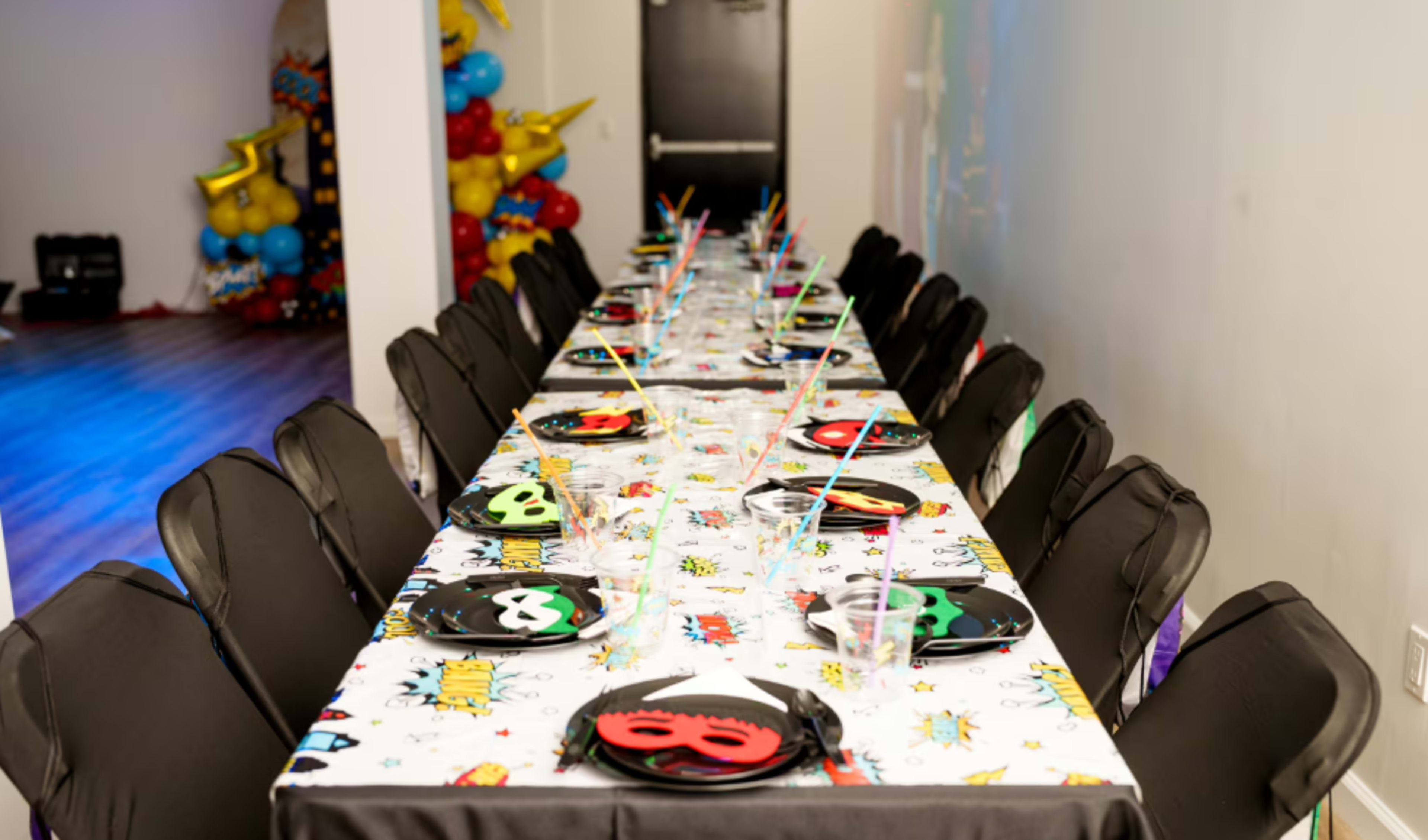 A long table is set with colorful party decorations, plates, masks, and straws in a room adorned with balloons.