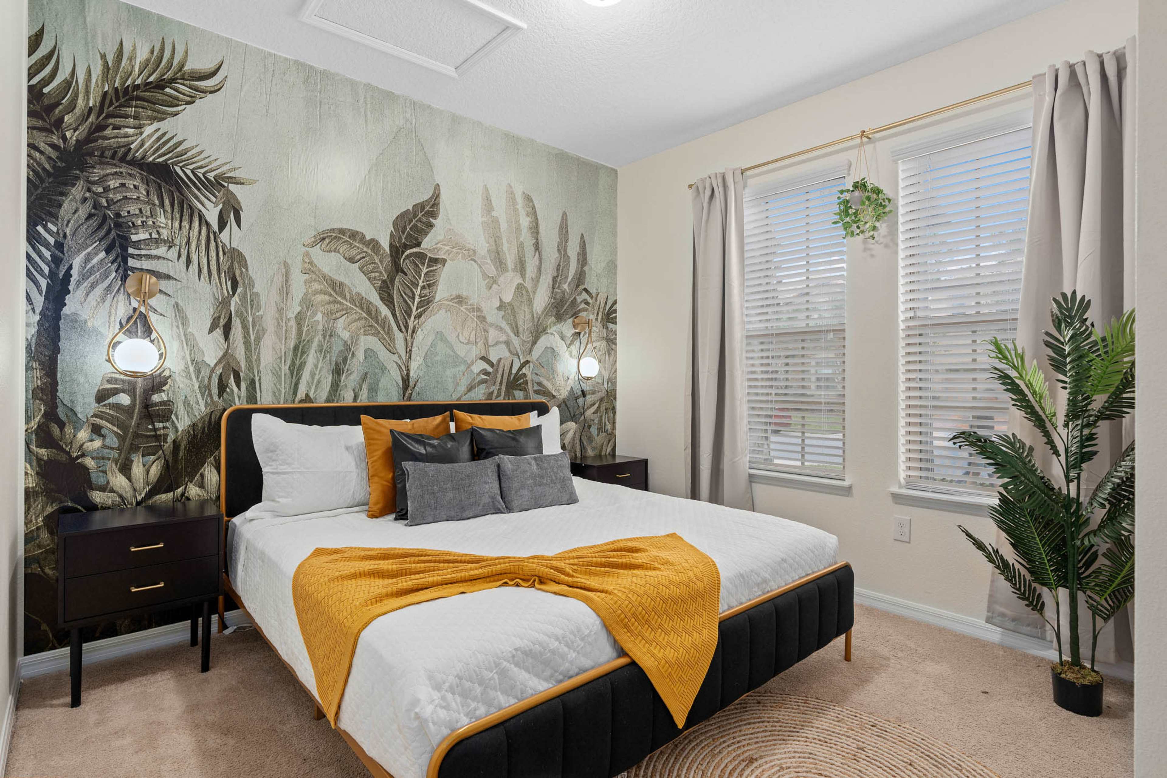 A bedroom features a large bed with an orange and gray throw blanket, flanked by two nightstands, and adorned with a tropical mural and potted plant.