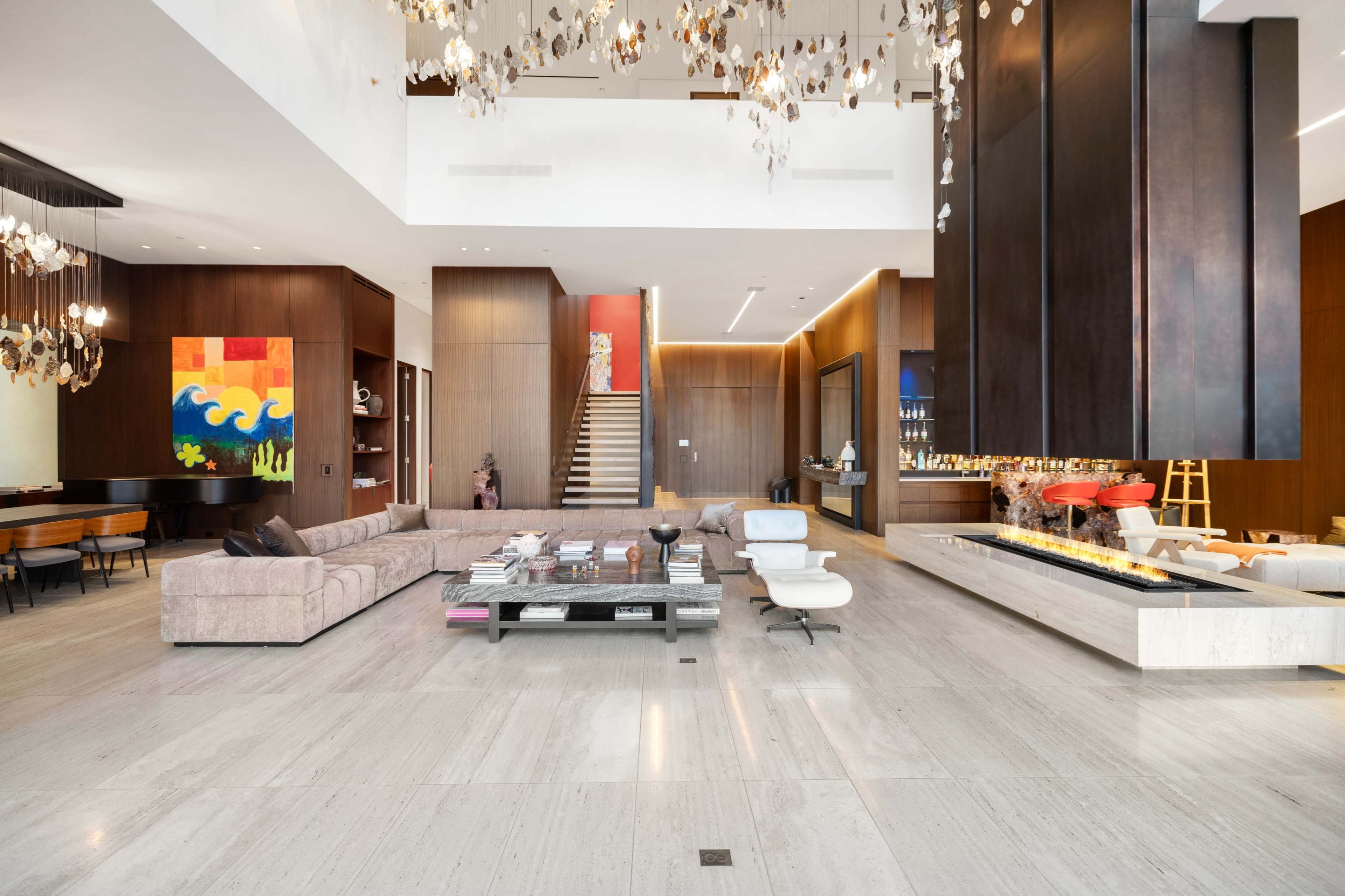 An expansive modern living room features a large sectional sofa, a sleek coffee table, and a striking fireplace, with a staircase and colorful artwork in the background.