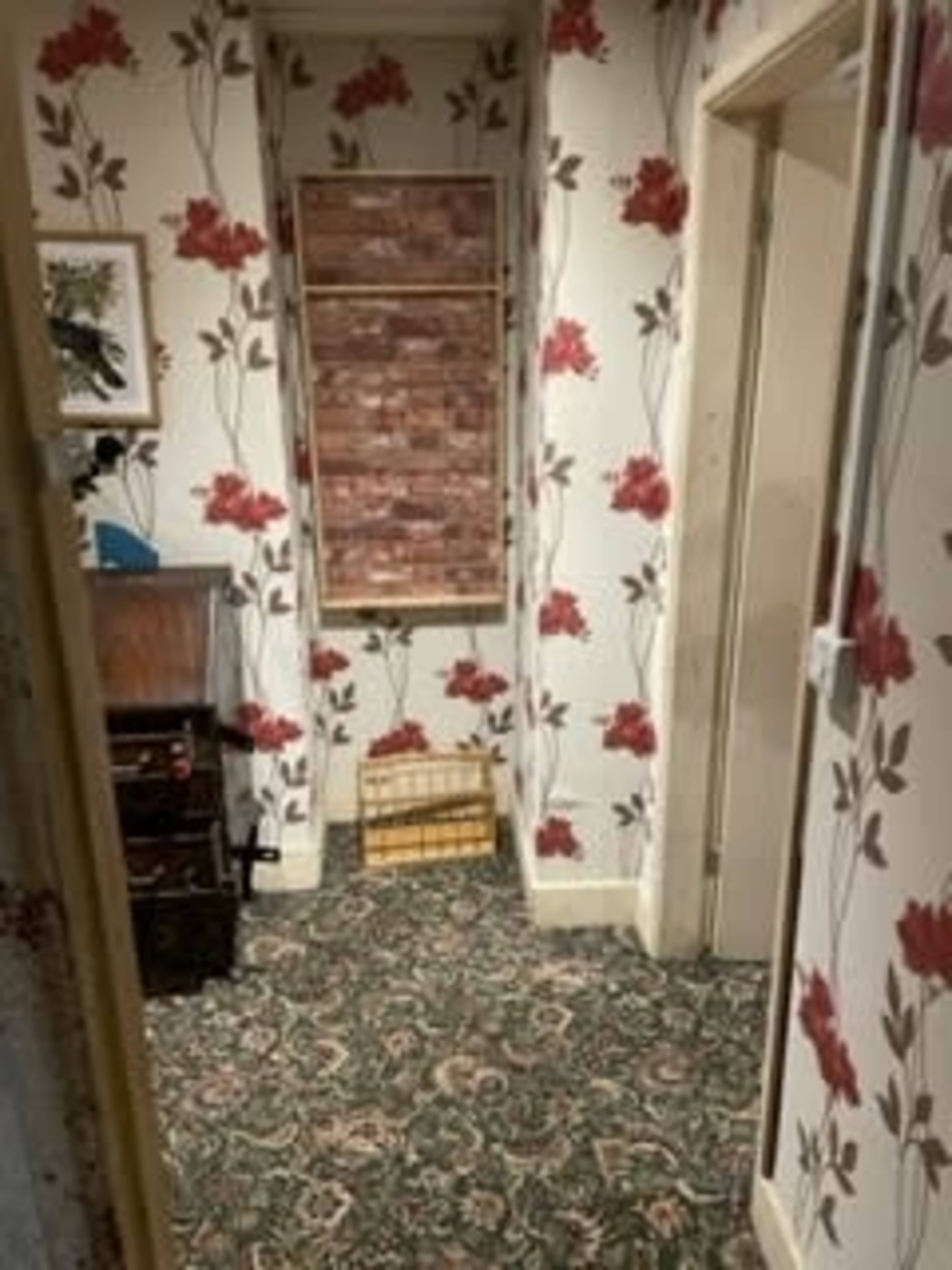 A narrow hallway with floral wallpaper, a patterned carpet, and a wooden chest of drawers near a partially closed window.