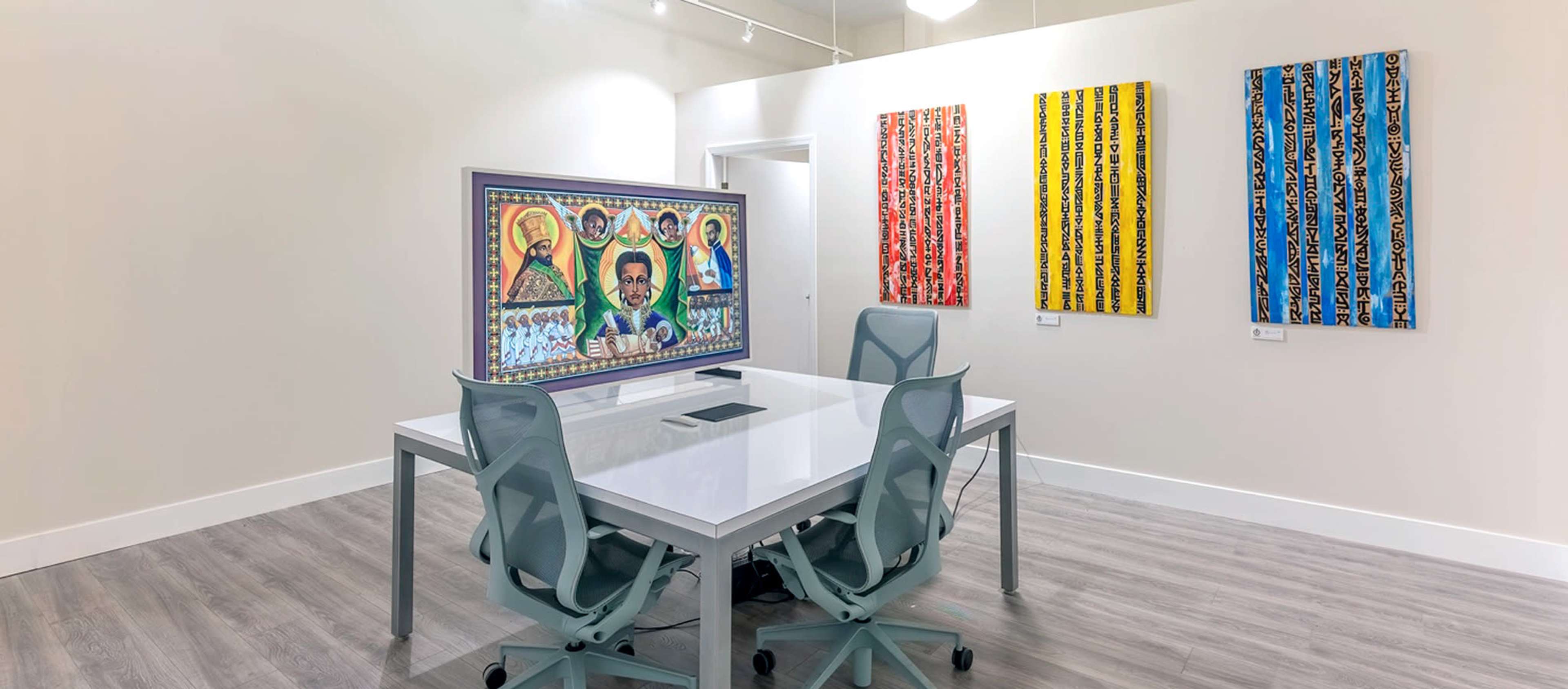 A modern art gallery featuring a white table and chairs in the center, with colorful paintings on the walls and a large screen displaying an artwork.