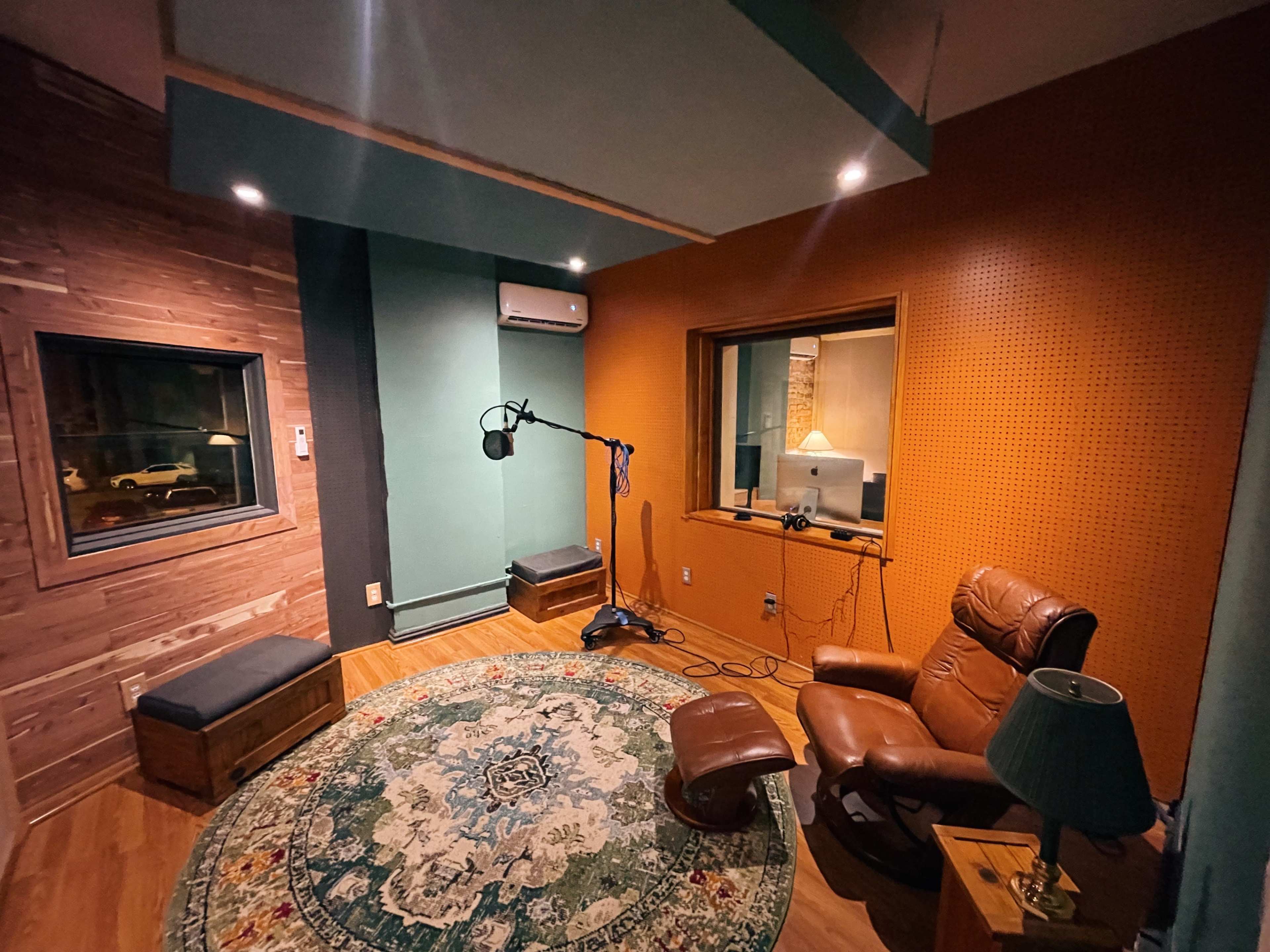 A professional recording studio with a microphone on a stand, a brown leather chair, and a large window overlooking a control room.