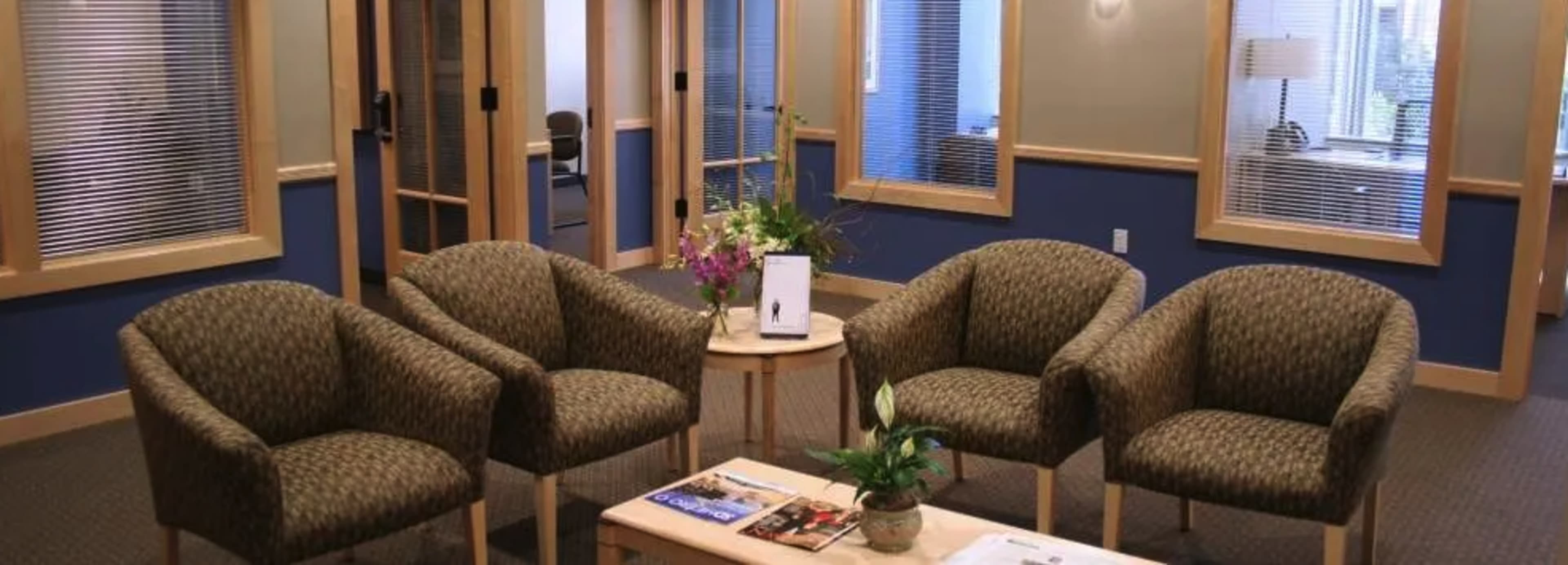 A waiting area with four patterned chairs arranged around a small wooden table featuring a plant and some magazines.