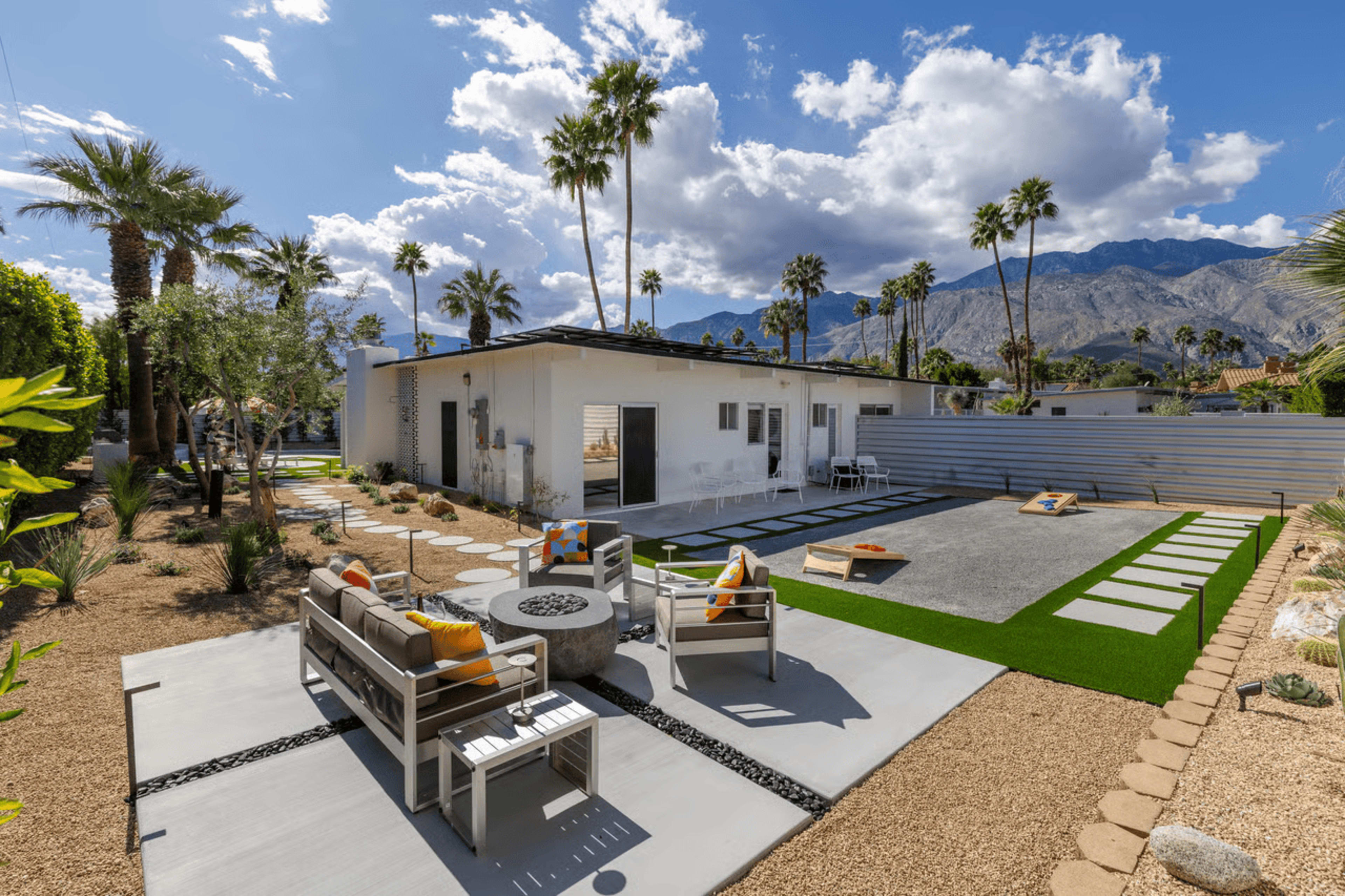 The image shows a modern residential backyard with a seating area, a fire pit, and a gravel space for games, surrounded by palm trees and mountains in the background.