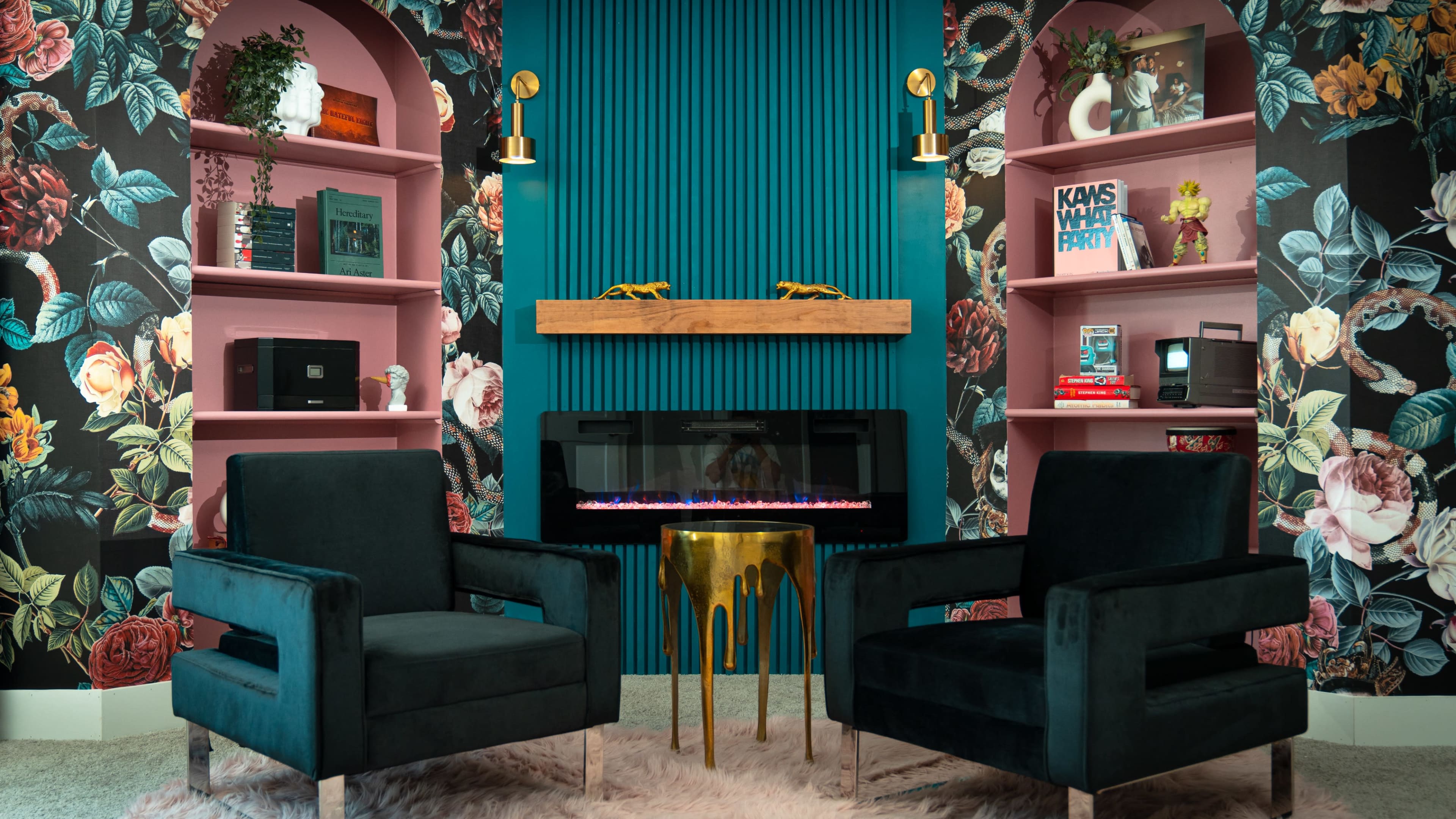 The image features a cozy seating area with two dark green chairs facing a modern electric fireplace, surrounded by floral-patterned wallpaper and pink shelves displaying various decorative items.