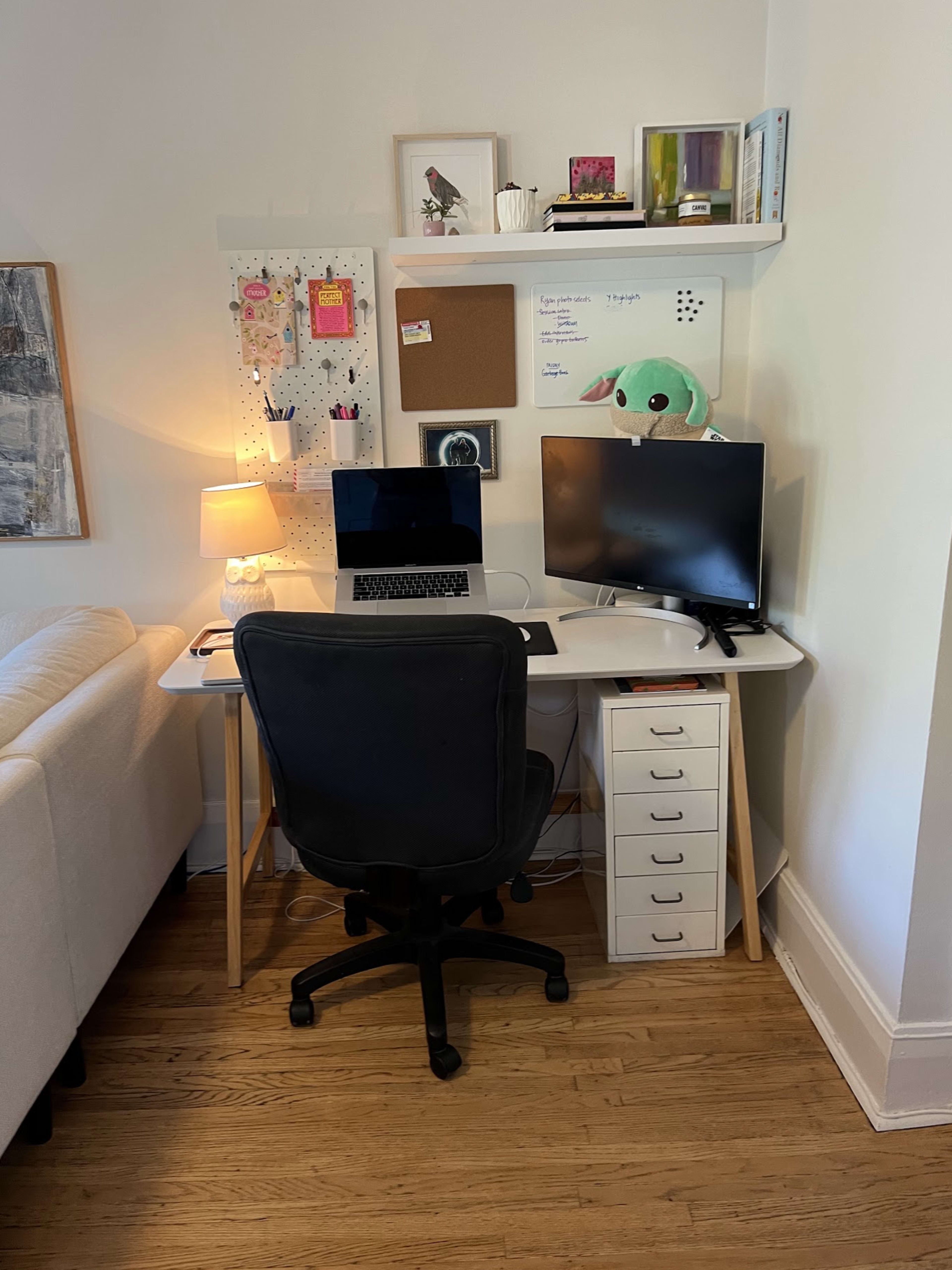A small home office setup features a desk with a laptop, a monitor, a lamp, and a cork board mounted on the wall, accompanied by a plush toy on the shelf above.